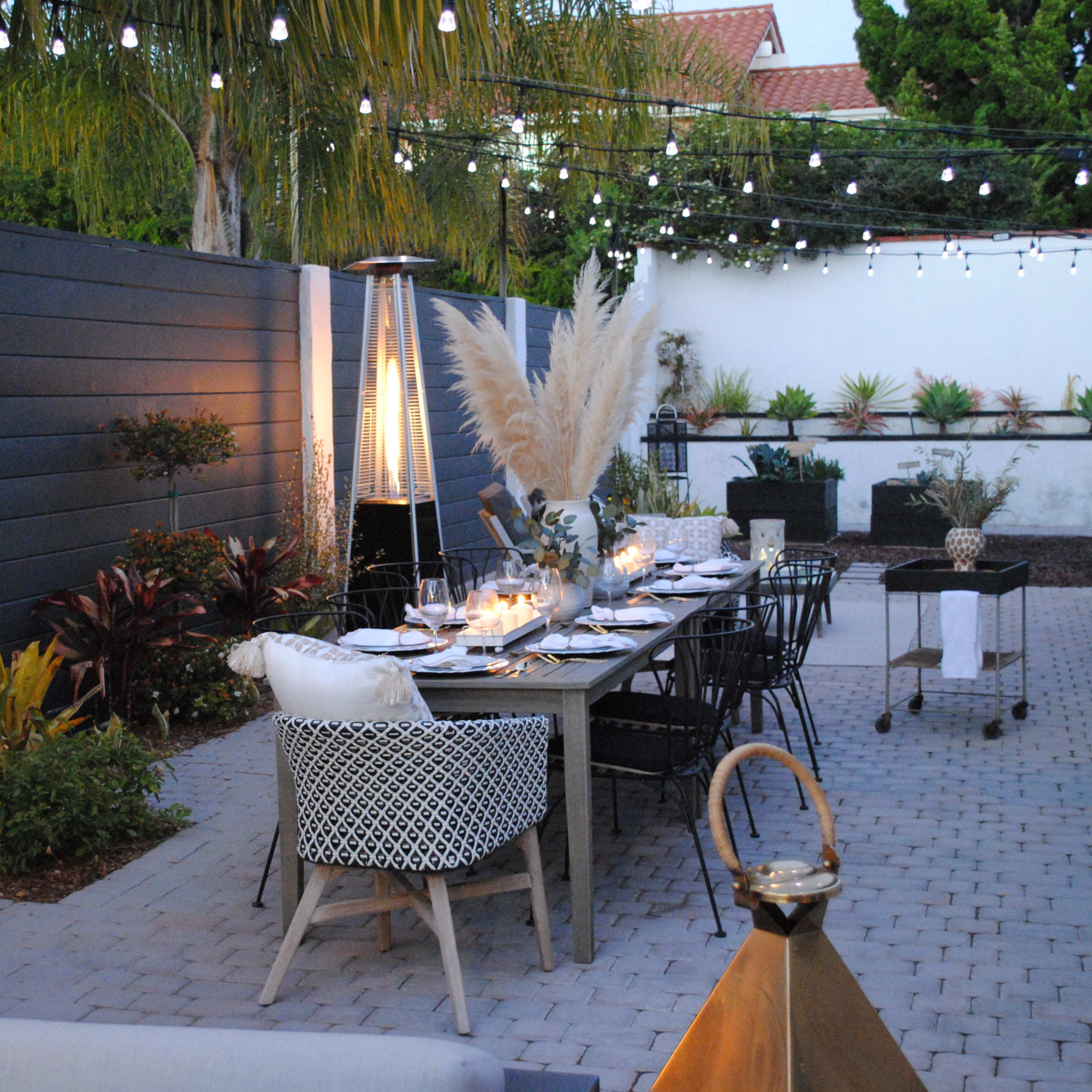 An outdoor dining area with a long table set for a meal, flanked by decorative plants and illuminated by string lights.
