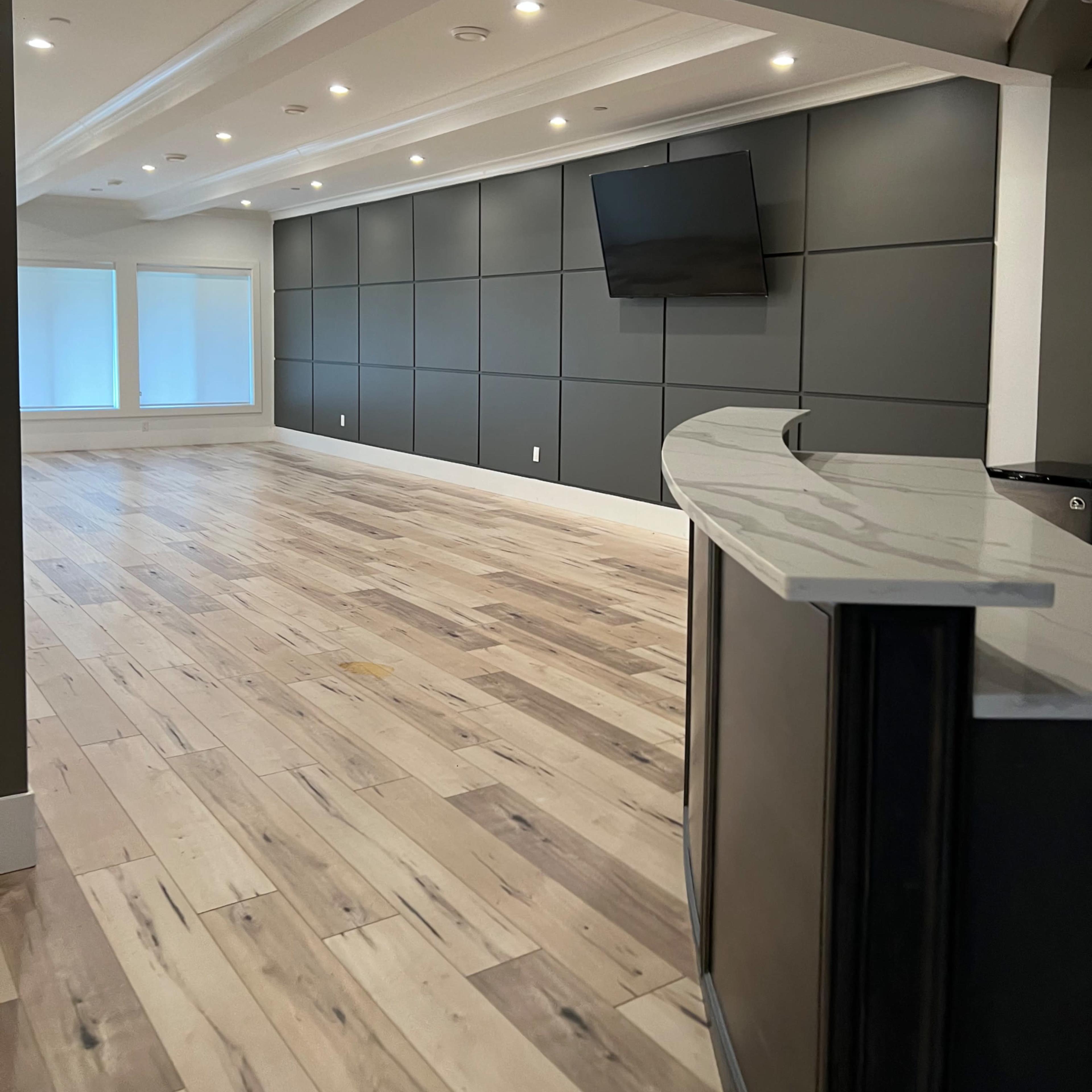 An empty room features a wood floor, a curved countertop, a mounted television on a dark paneled wall, and two windows with blinds.