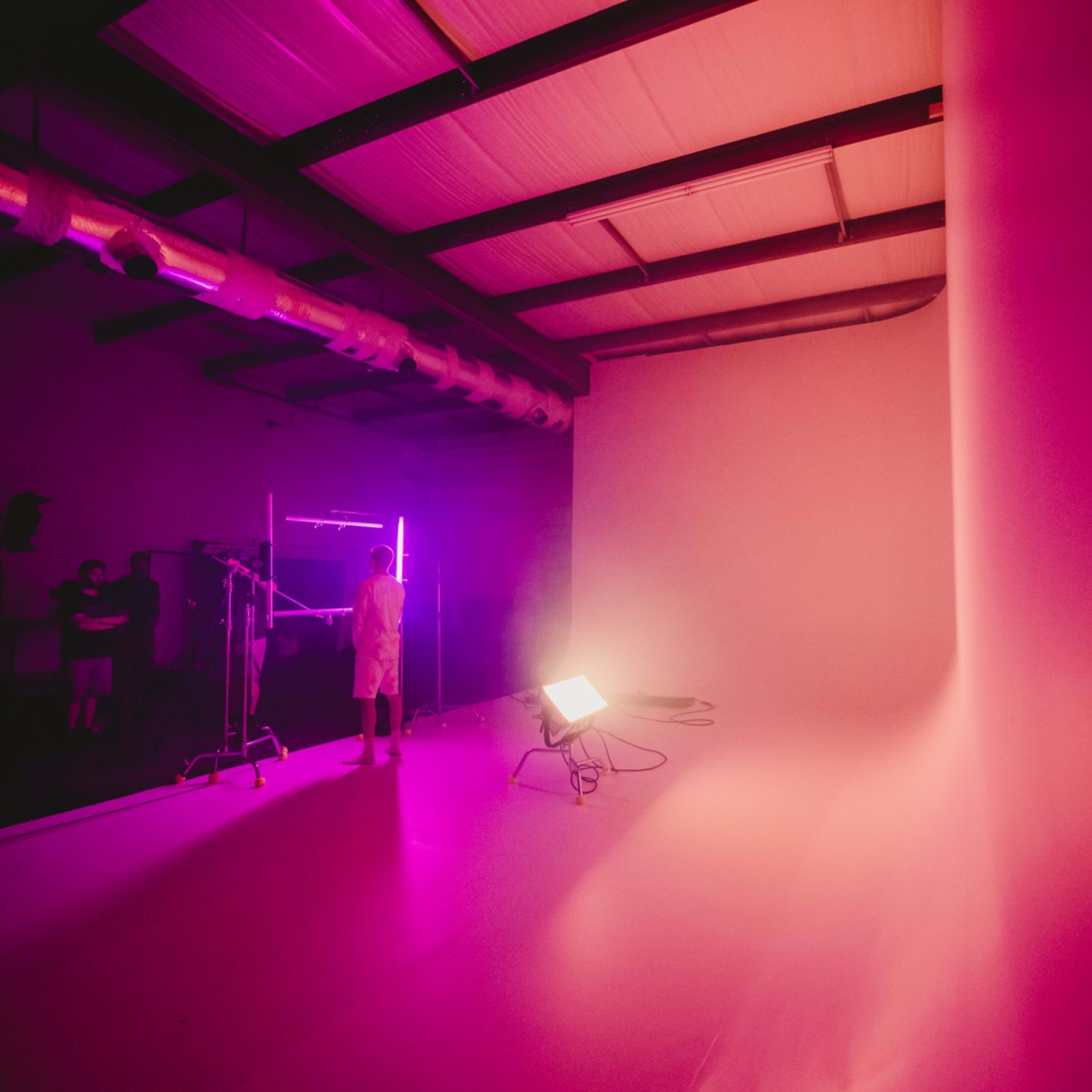 A person stands in a photography studio illuminated by pink and purple lights, with a seamless backdrop and equipment in the background.