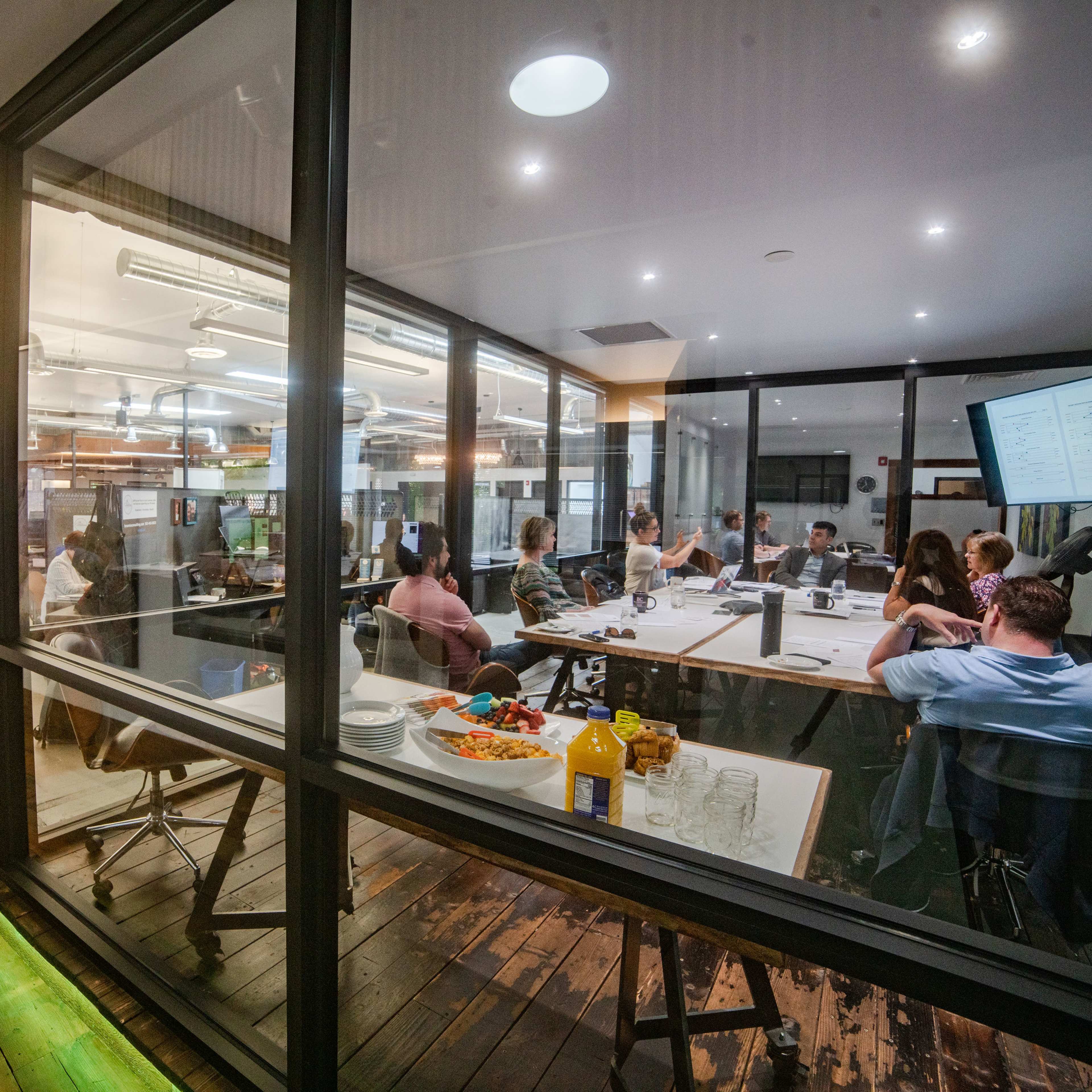 A group of people is engaged in a meeting in a glass-walled conference room, with snacks visible on a nearby table.