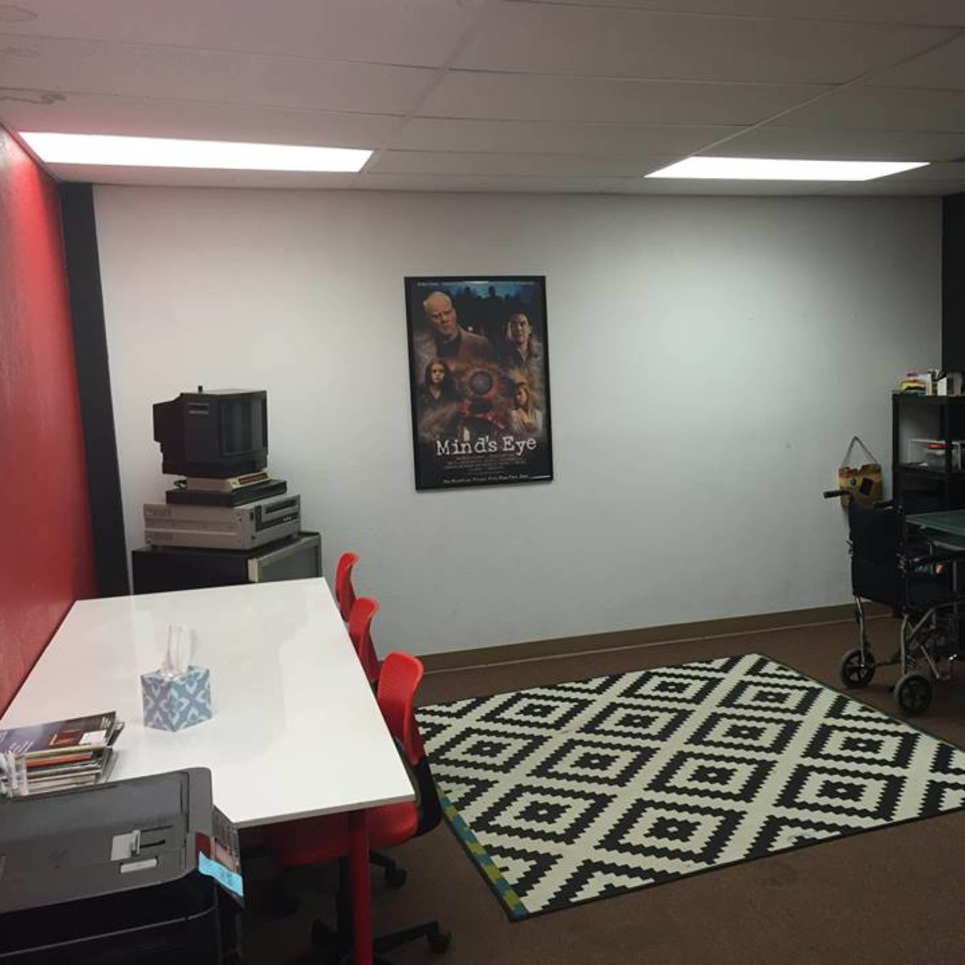 A sparsely furnished room with a table and red chairs, a retro television, an arcade cabinet, and a poster on the wall.