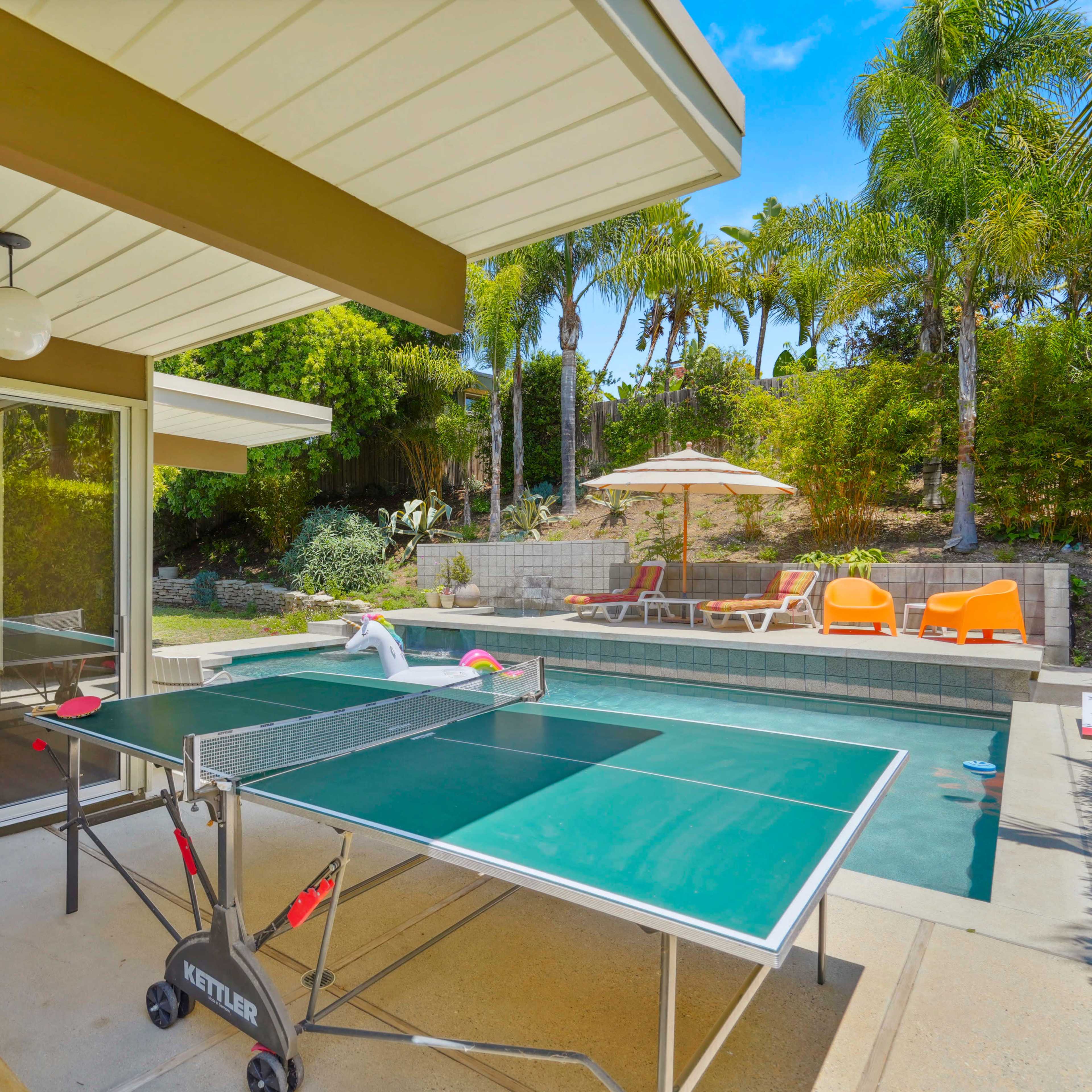 A ping pong table is set up on a patio beside a pool, surrounded by palm trees and lounge chairs.