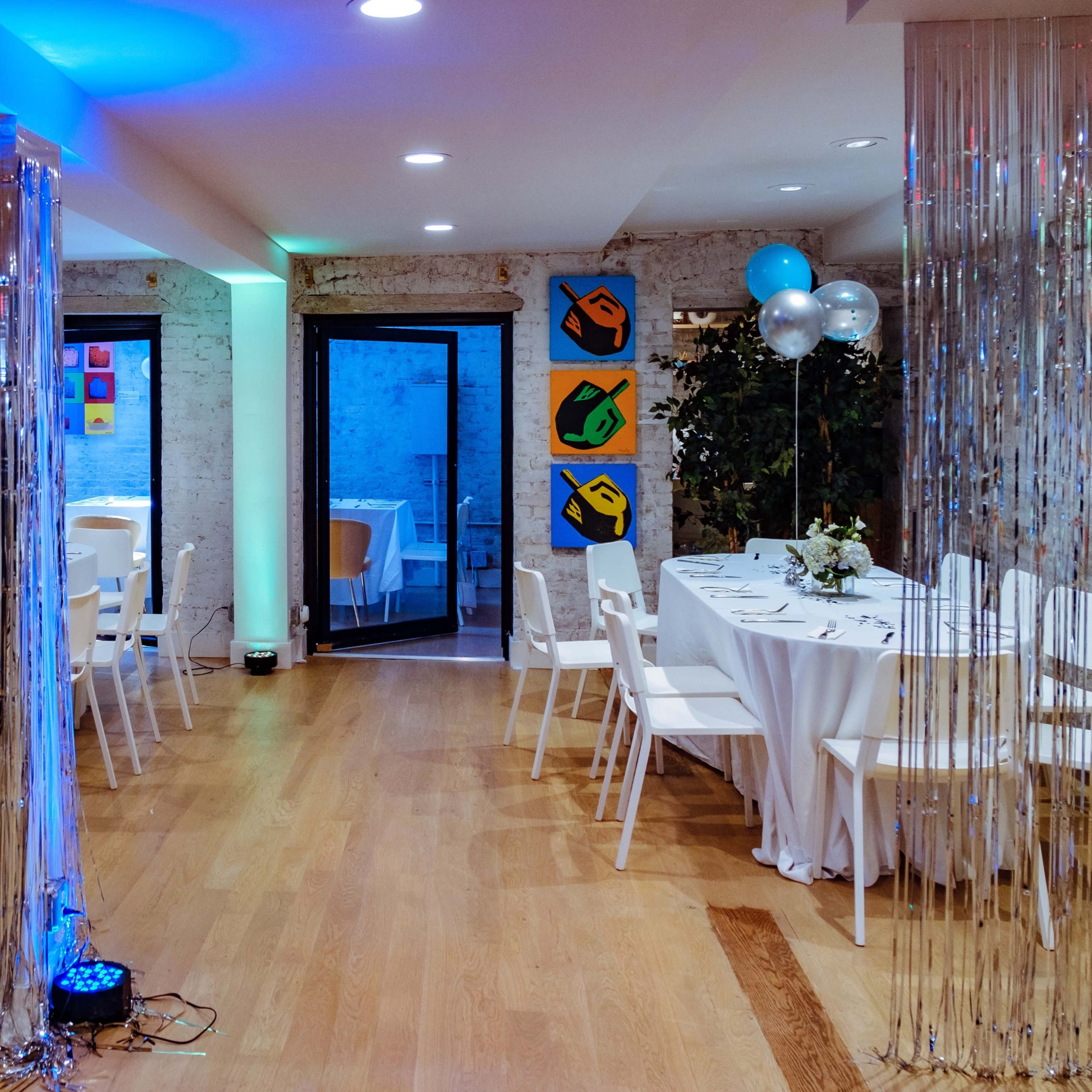 A dining space features round tables set with white tablecloths, illuminated by blue and silver decorations, and adjacent to colorful artwork on the walls.