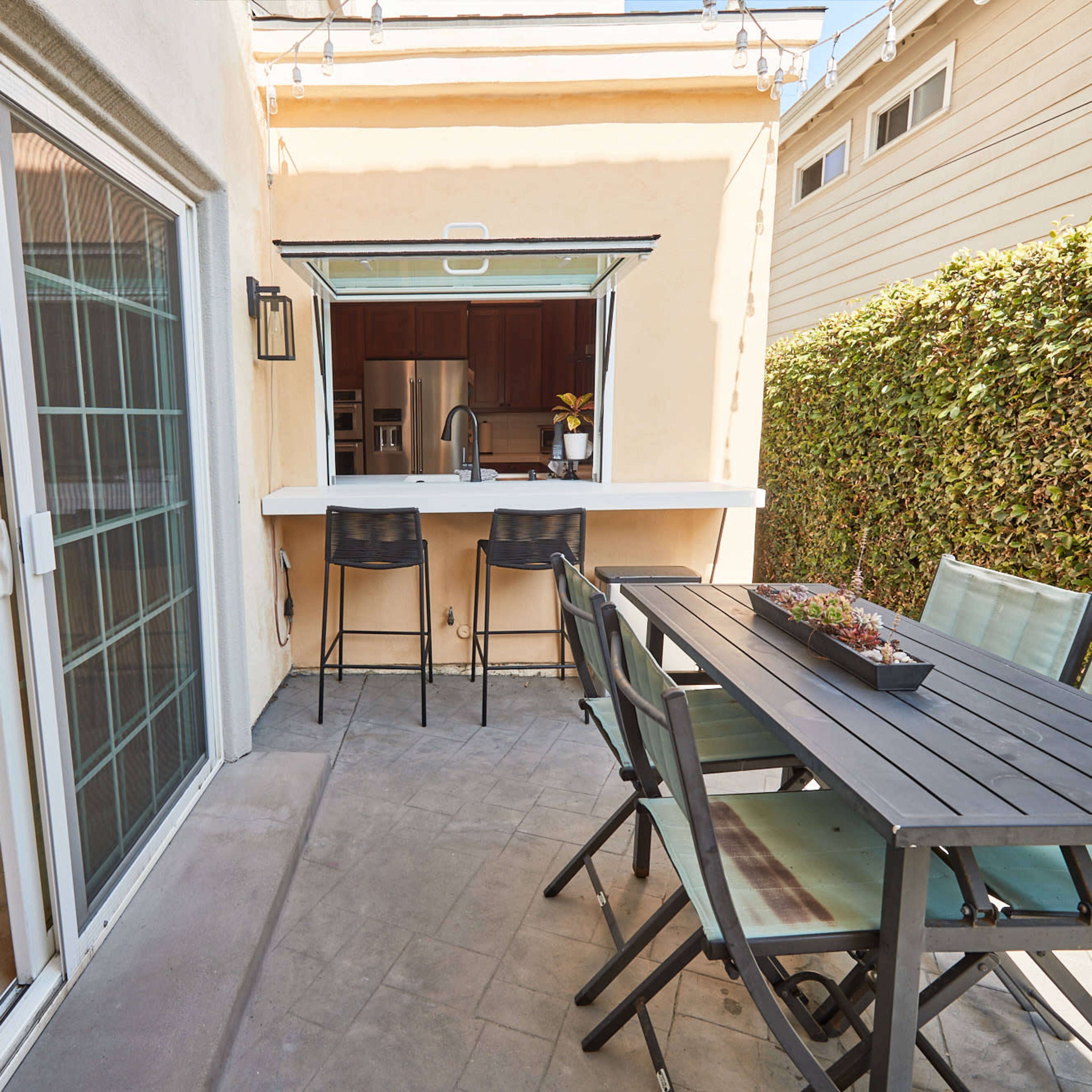 A patio area features a dining table with chairs, a bar counter with stools, and a view of an adjacent kitchen through a window.