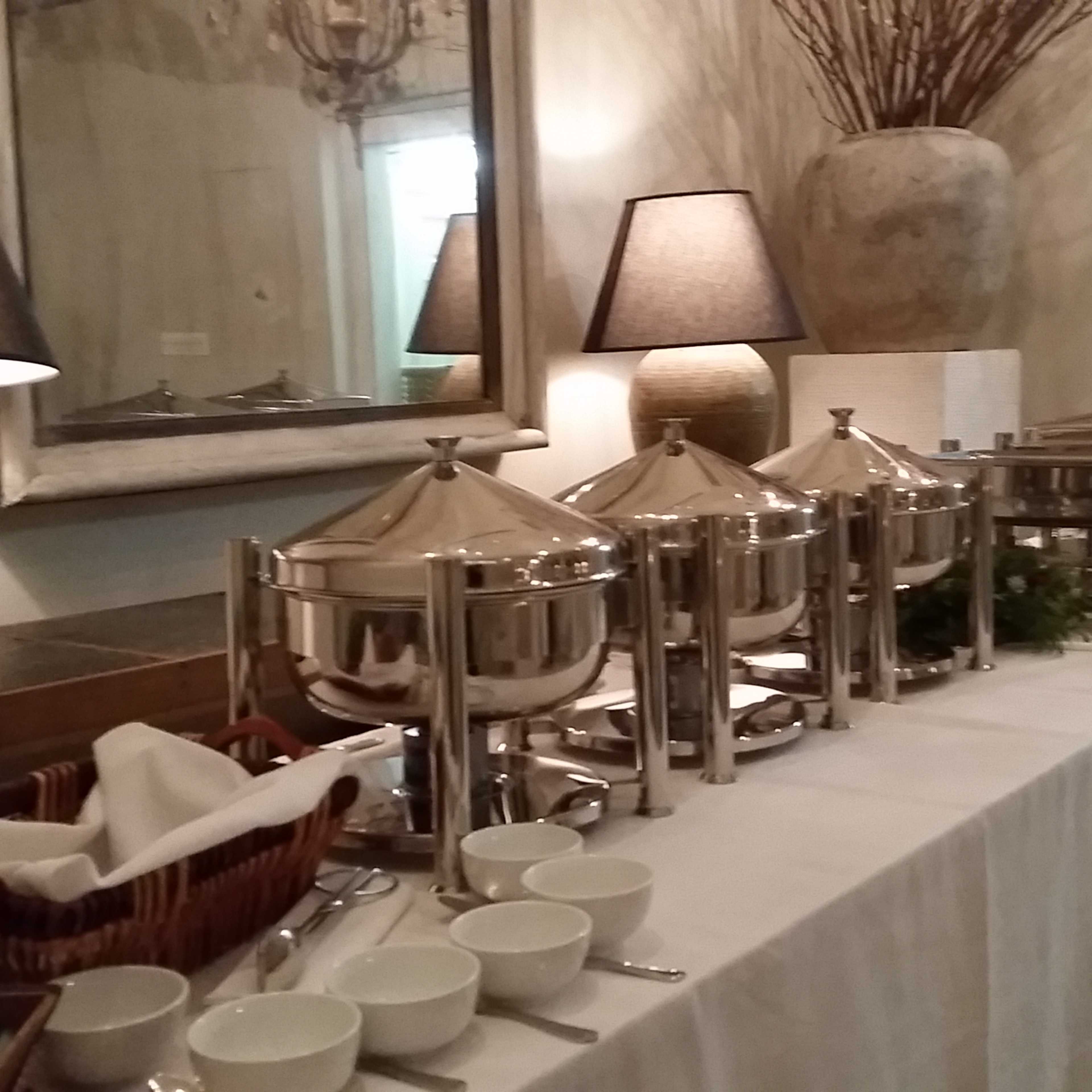 A long table is set with covered food trays, bowls, and decorative lamps.