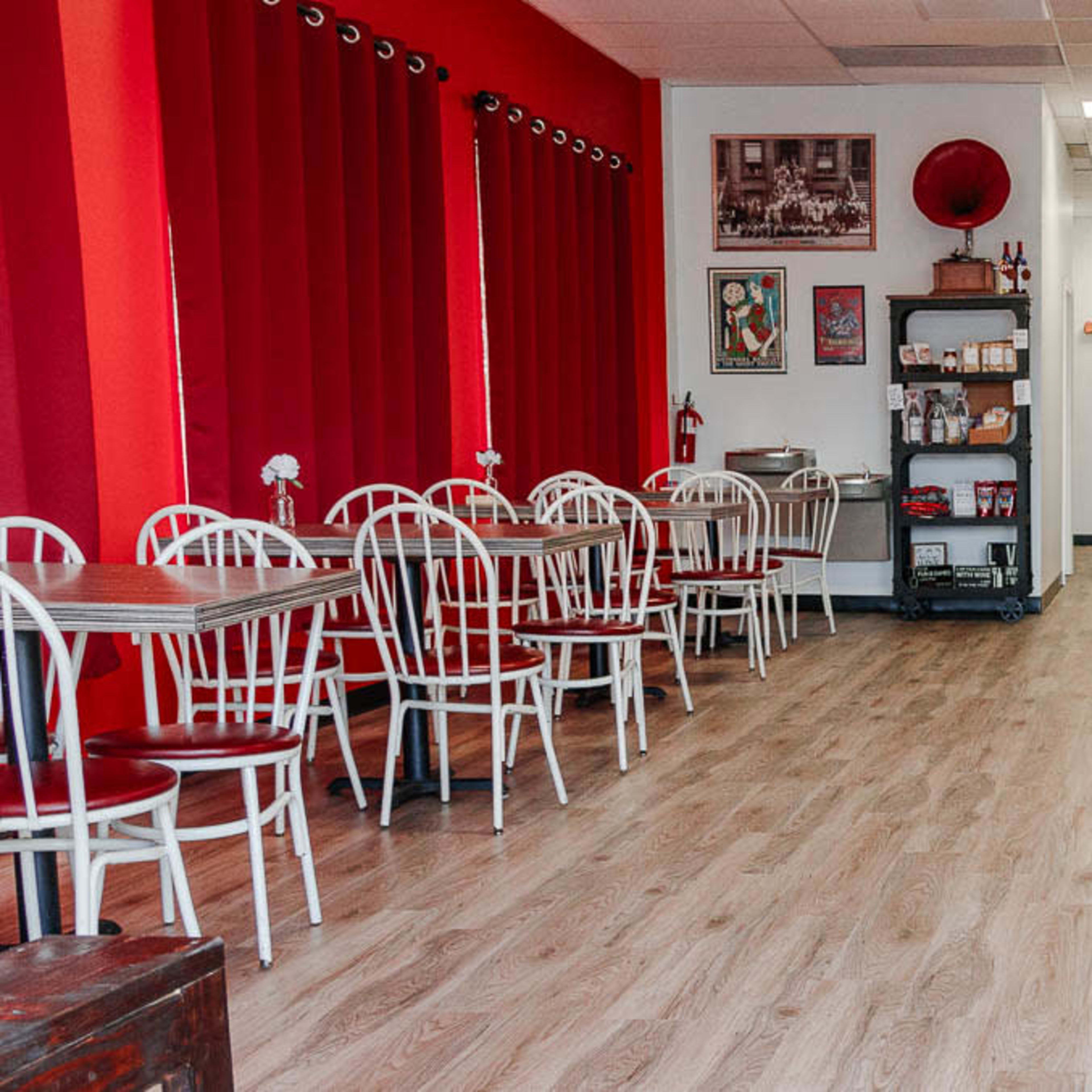 A café interior with red curtains, white tables and chairs, and a wooden counter along one side.