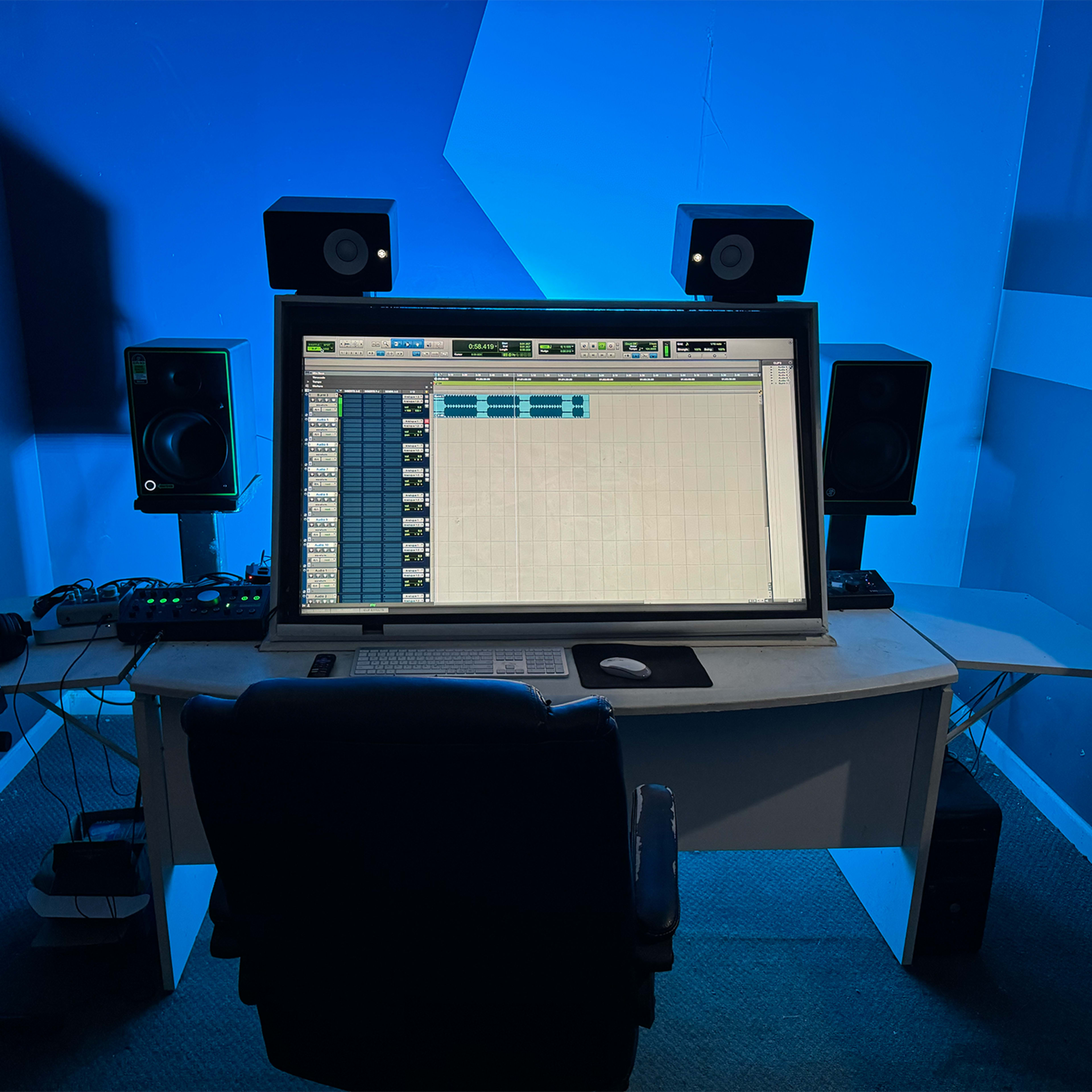 A music production studio features a large computer monitor displaying audio software, flanked by studio monitors and a keyboard in a blue-lit environment.