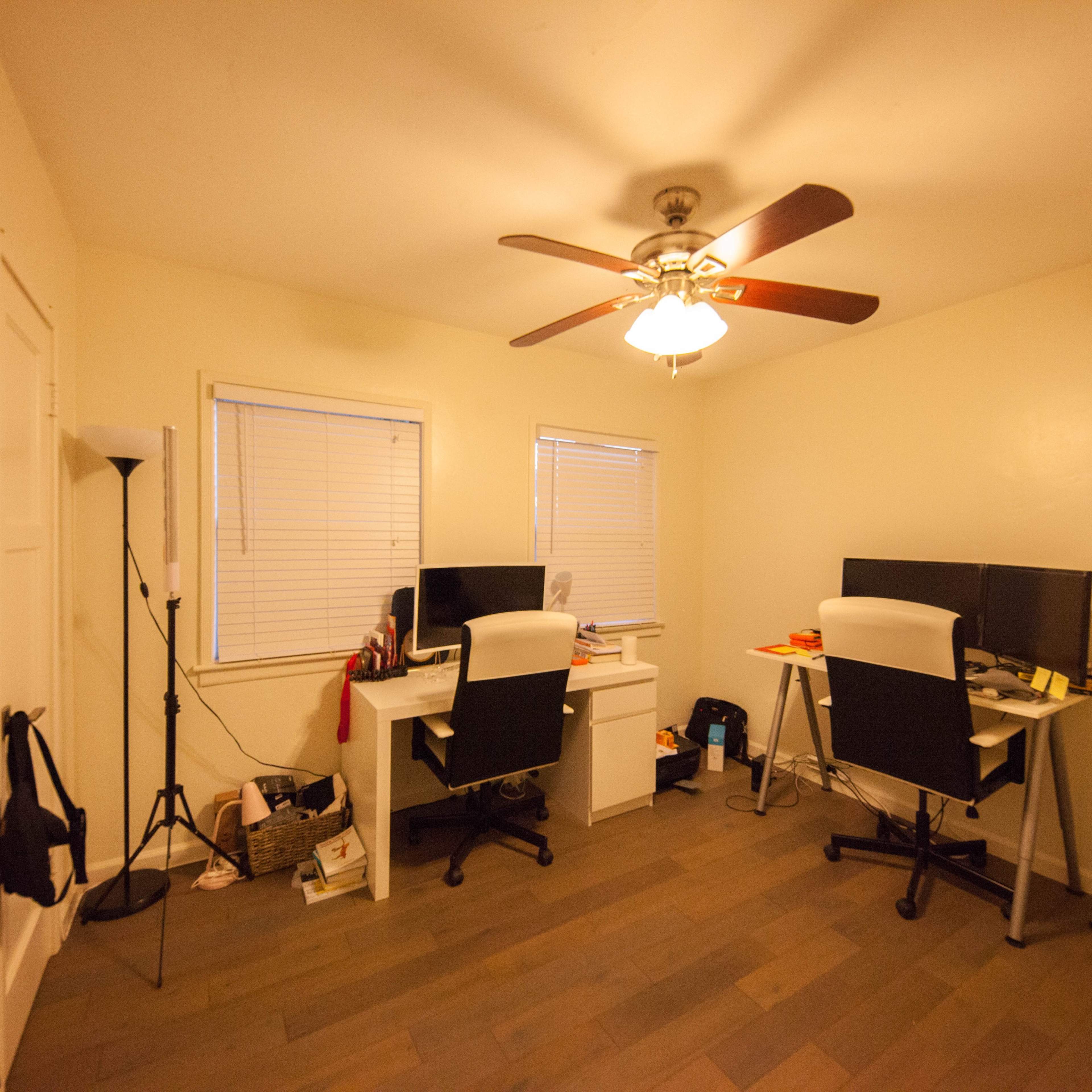 A small office space with two workstations, each featuring a computer and a chair, illuminated by a ceiling fan.