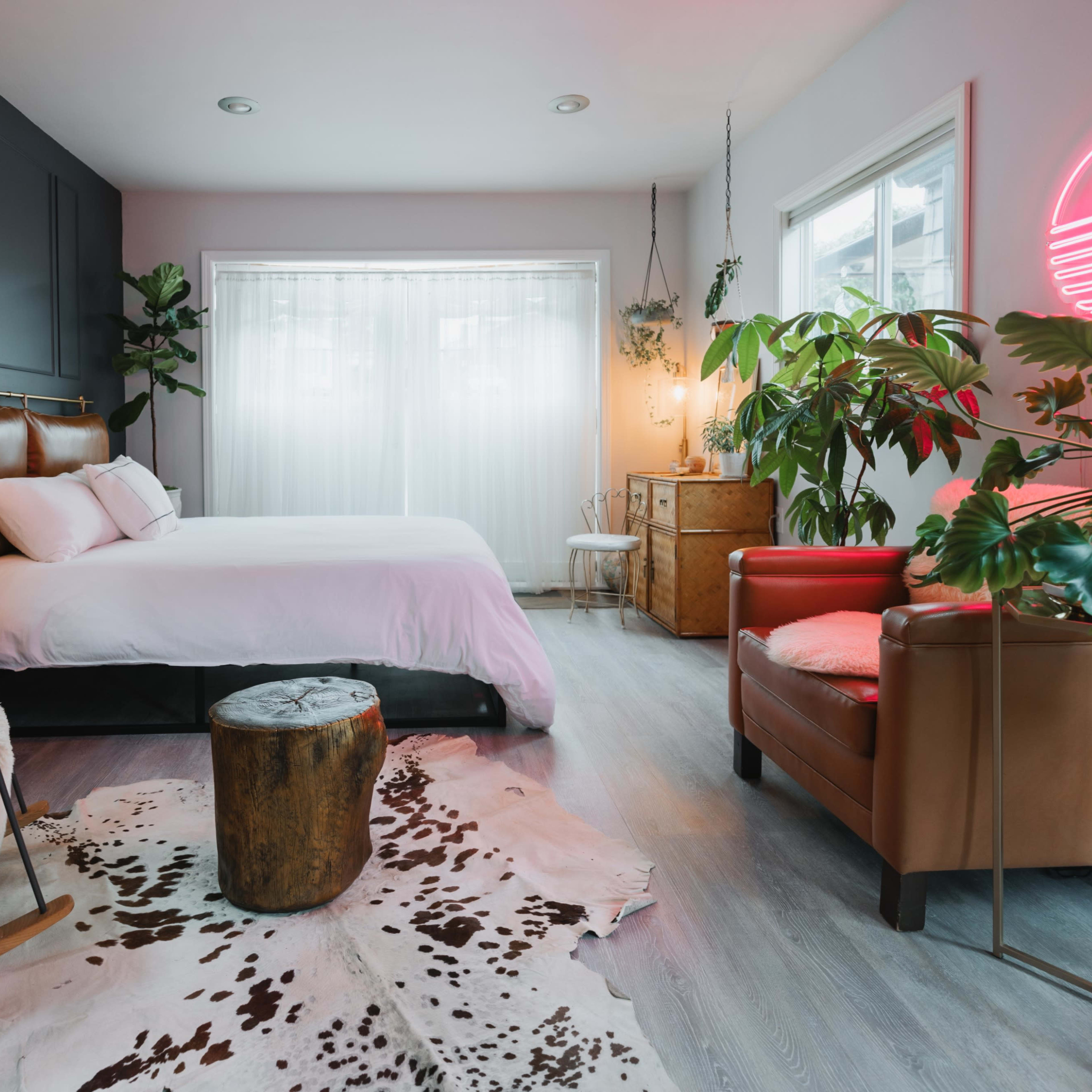 A modern bedroom featuring a bed with a brown leather headboard, a wooden rocking chair, a potted plant, and a neon light fixture.