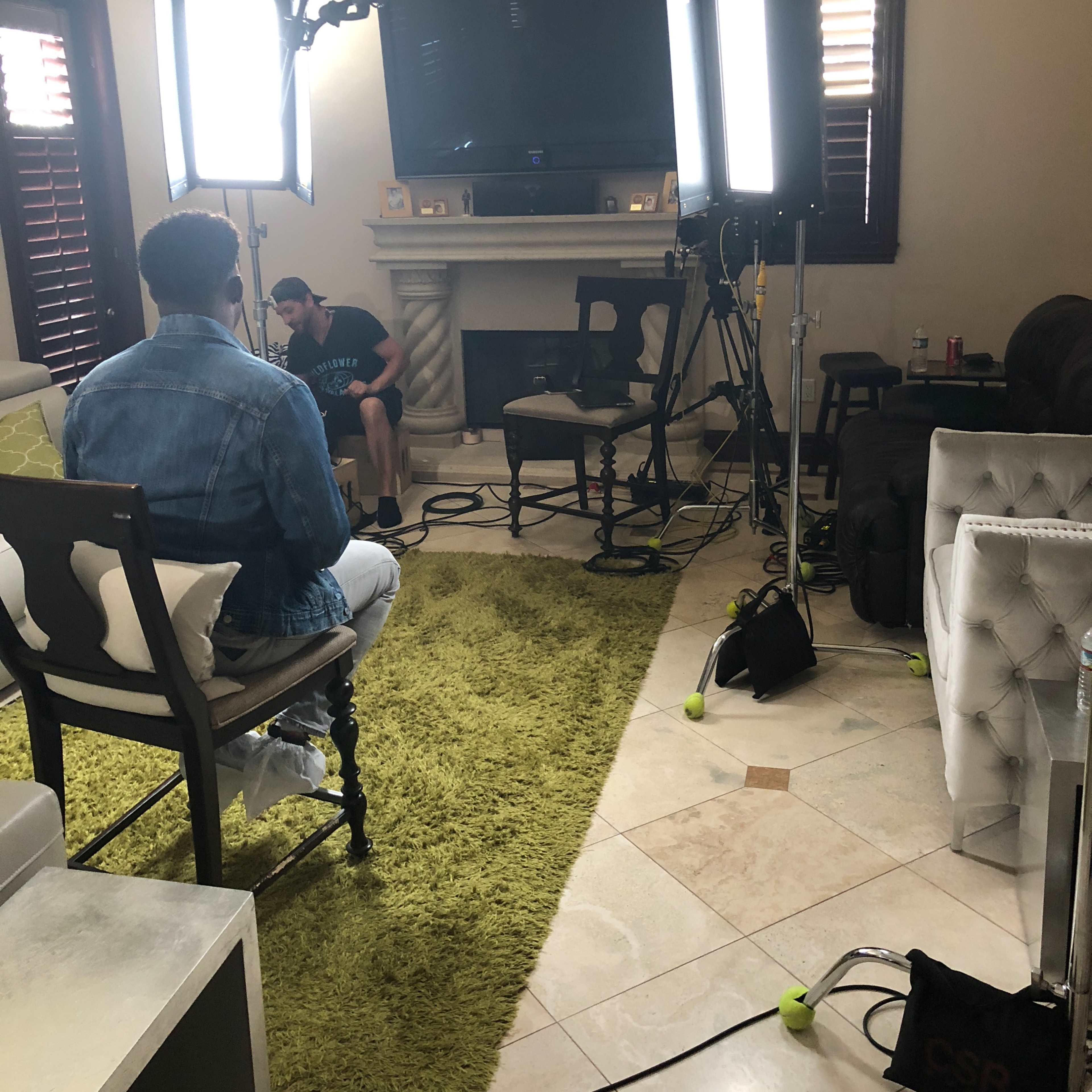A living room is set up for a video shoot, featuring bright lights, a camera, and a person sitting in front of a television.