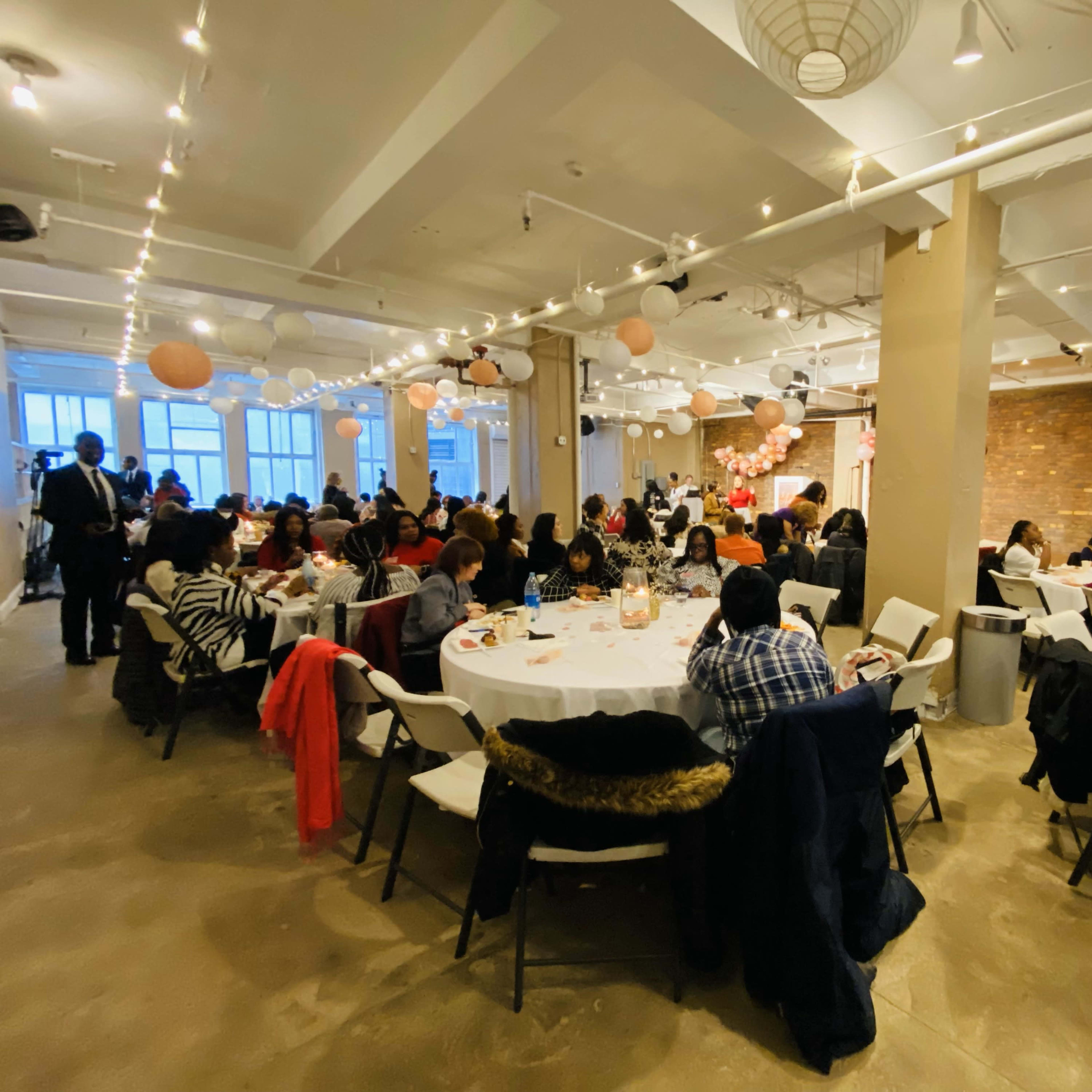 A large indoor event space is filled with round tables where many people are seated, dining and socializing under decorative lighting and hanging lanterns.
