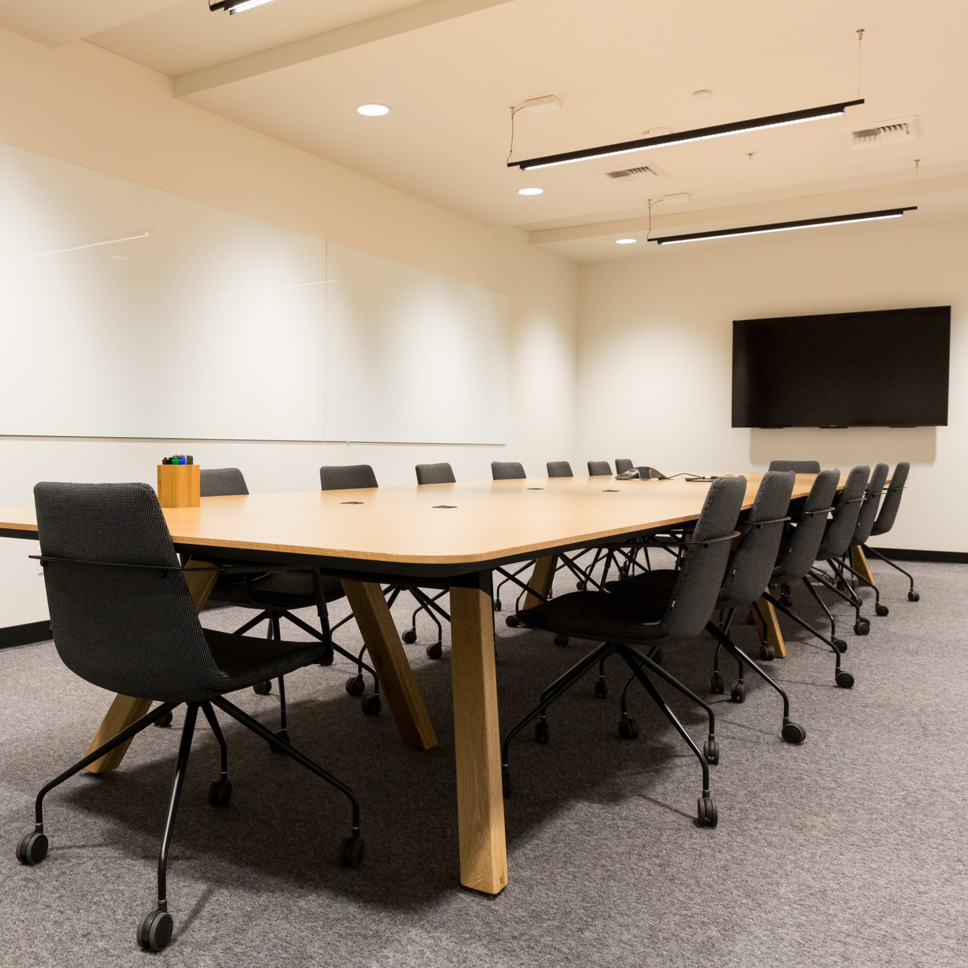 A large conference room features a long wooden table surrounded by black rolling chairs and a wall-mounted television.