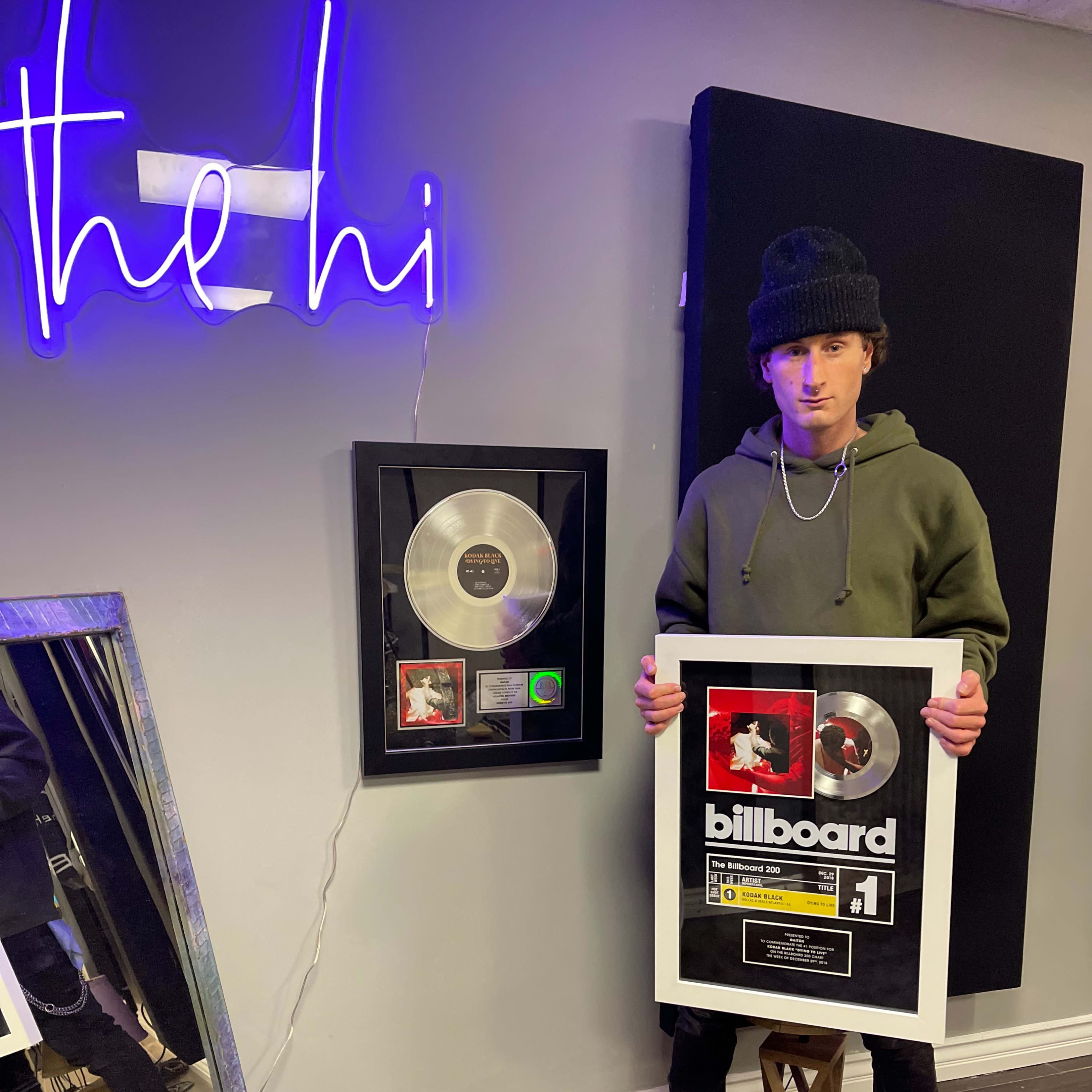A person wearing a green hoodie and beanie stands next to a framed silver record plaque and a neon sign that says "the hi."