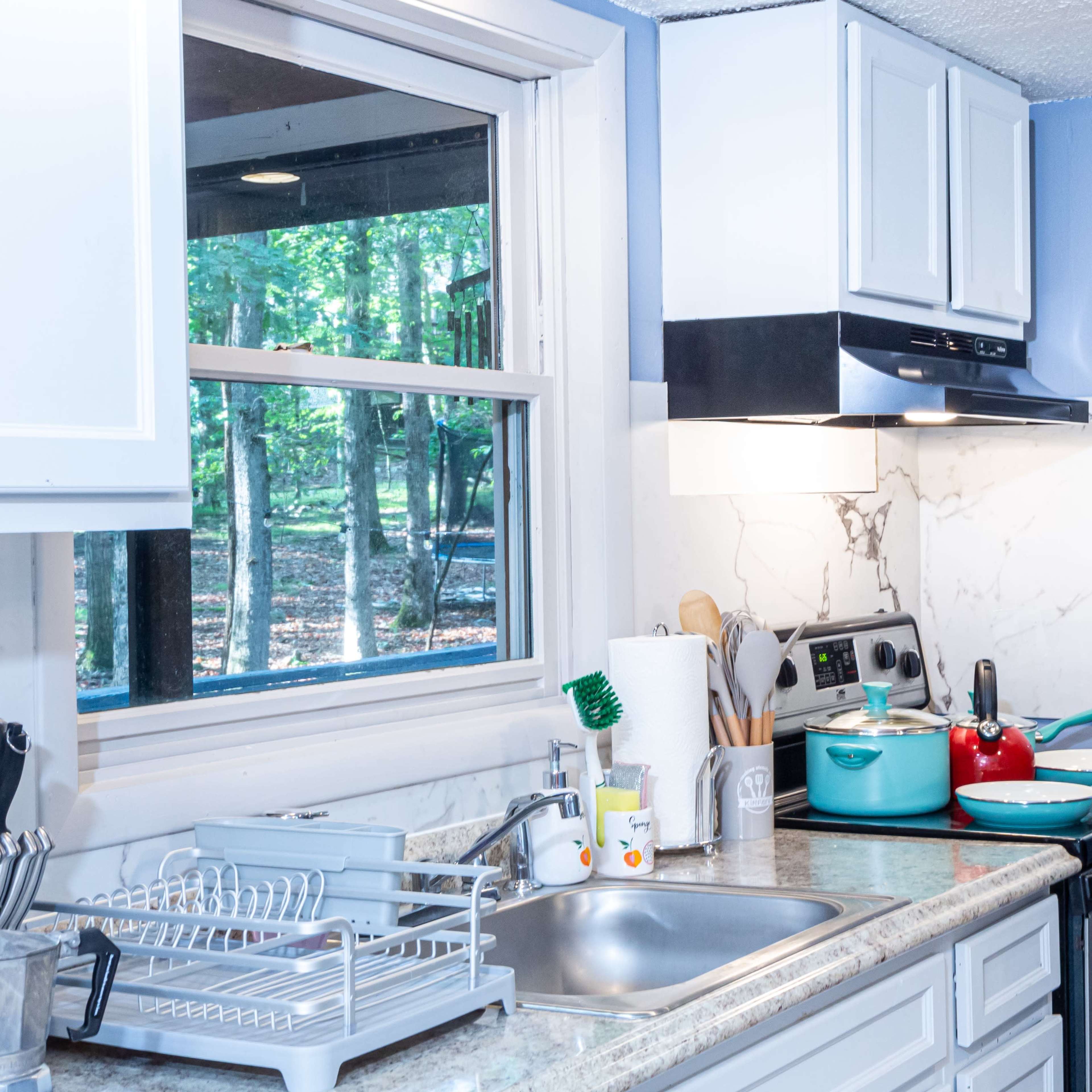 A kitchen with a sink, stove, and various kitchen utensils on a countertop, beside a window overlooking a wooded area.