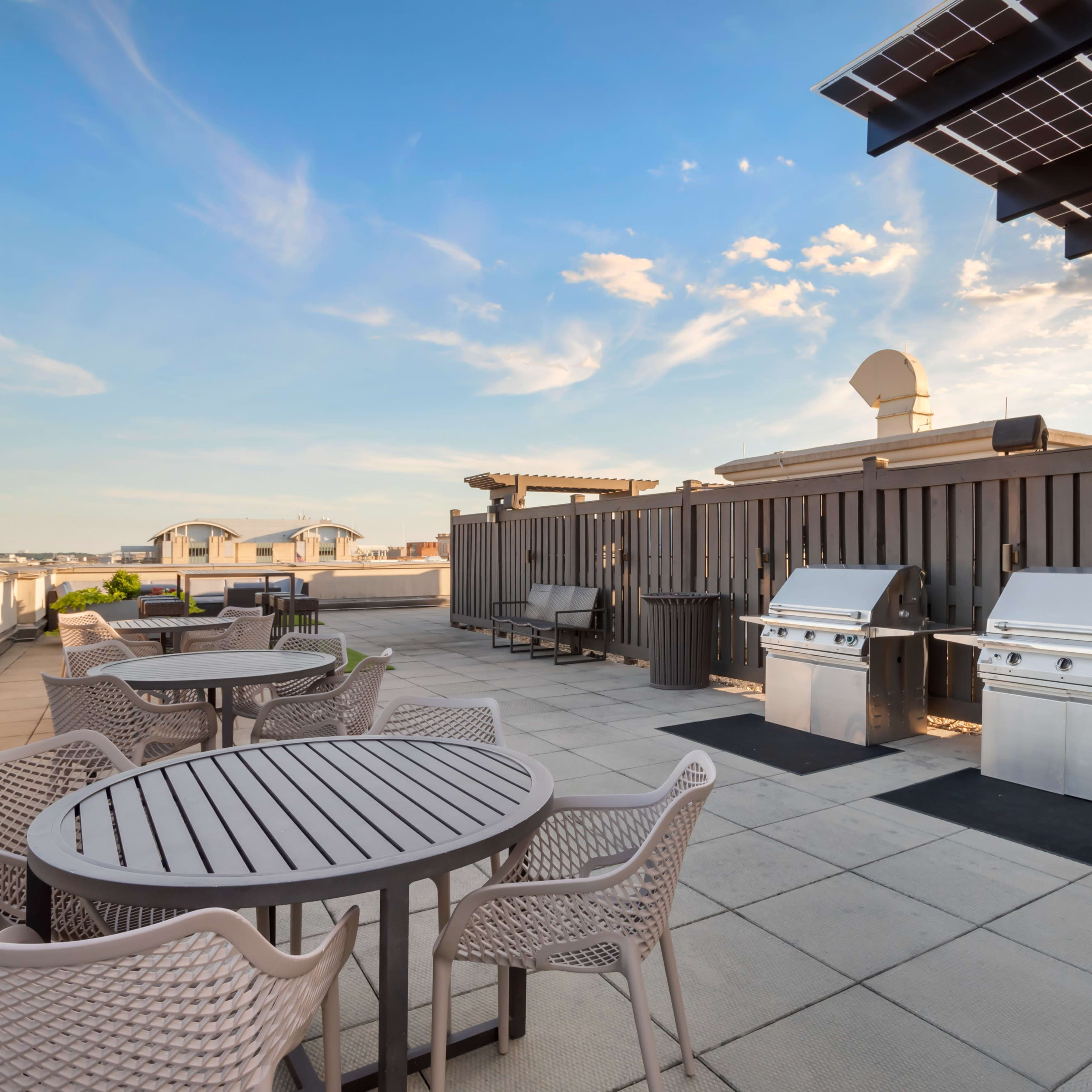A rooftop space featuring two stainless steel grills, a set of round tables with chairs, and solar panels overhead against a clear blue sky.