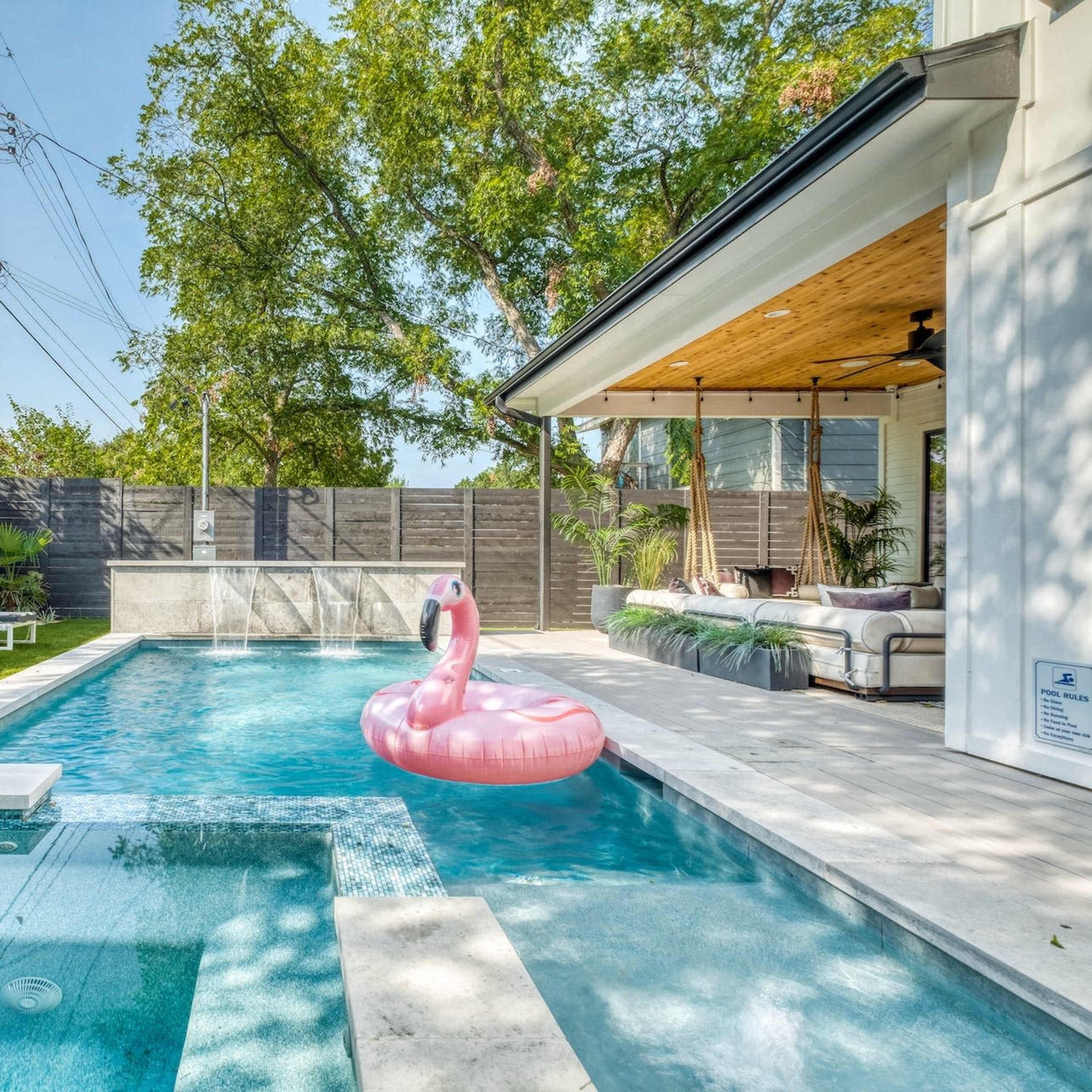 A pink flamingo float sits in a blue swimming pool surrounded by lounge chairs and landscaped greenery.