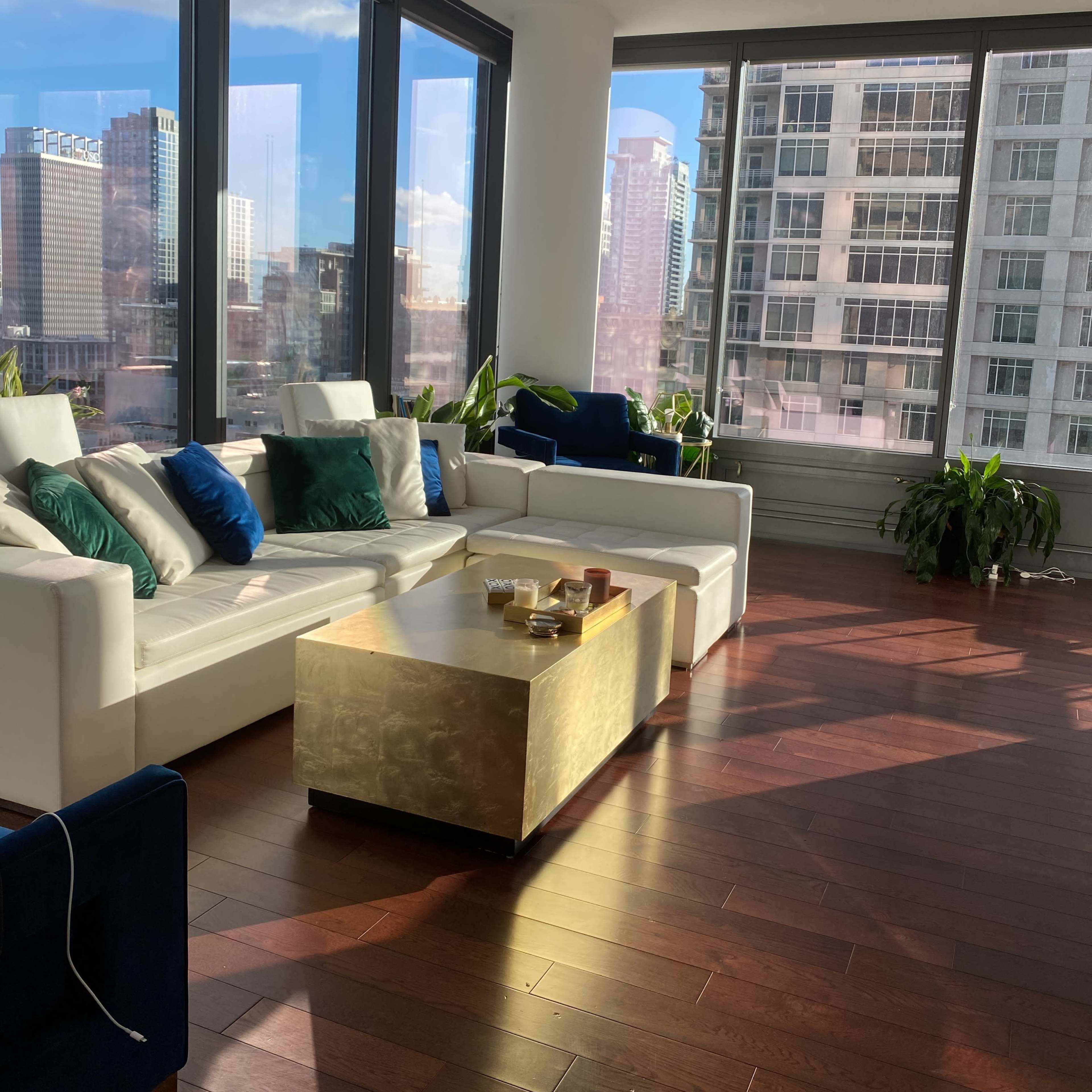 A spacious living area features a white sofa with blue and green cushions, a gold coffee table, and large windows displaying a city skyline.