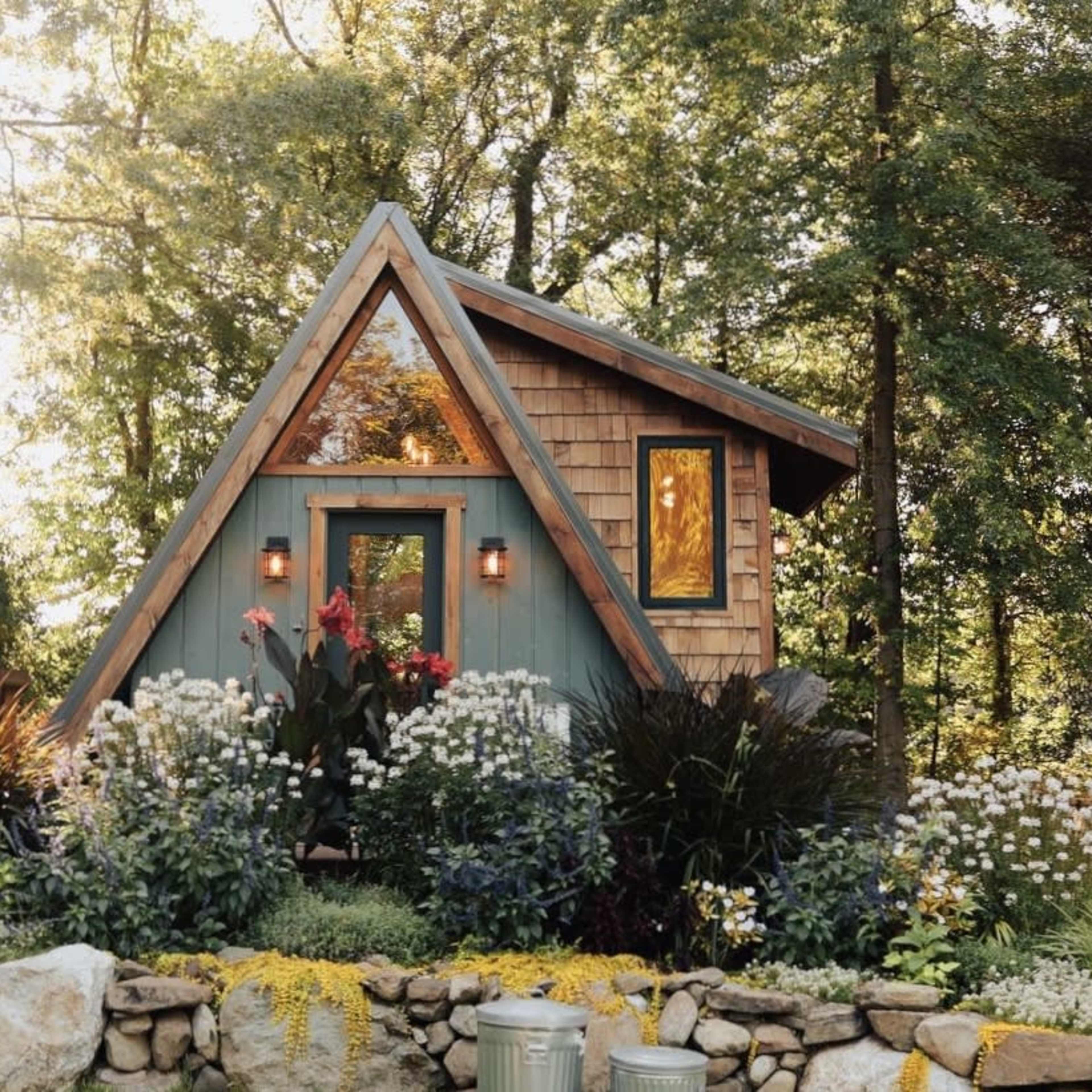 A small A-frame cabin nestled among trees, surrounded by a landscaped garden with various flowers and plants.