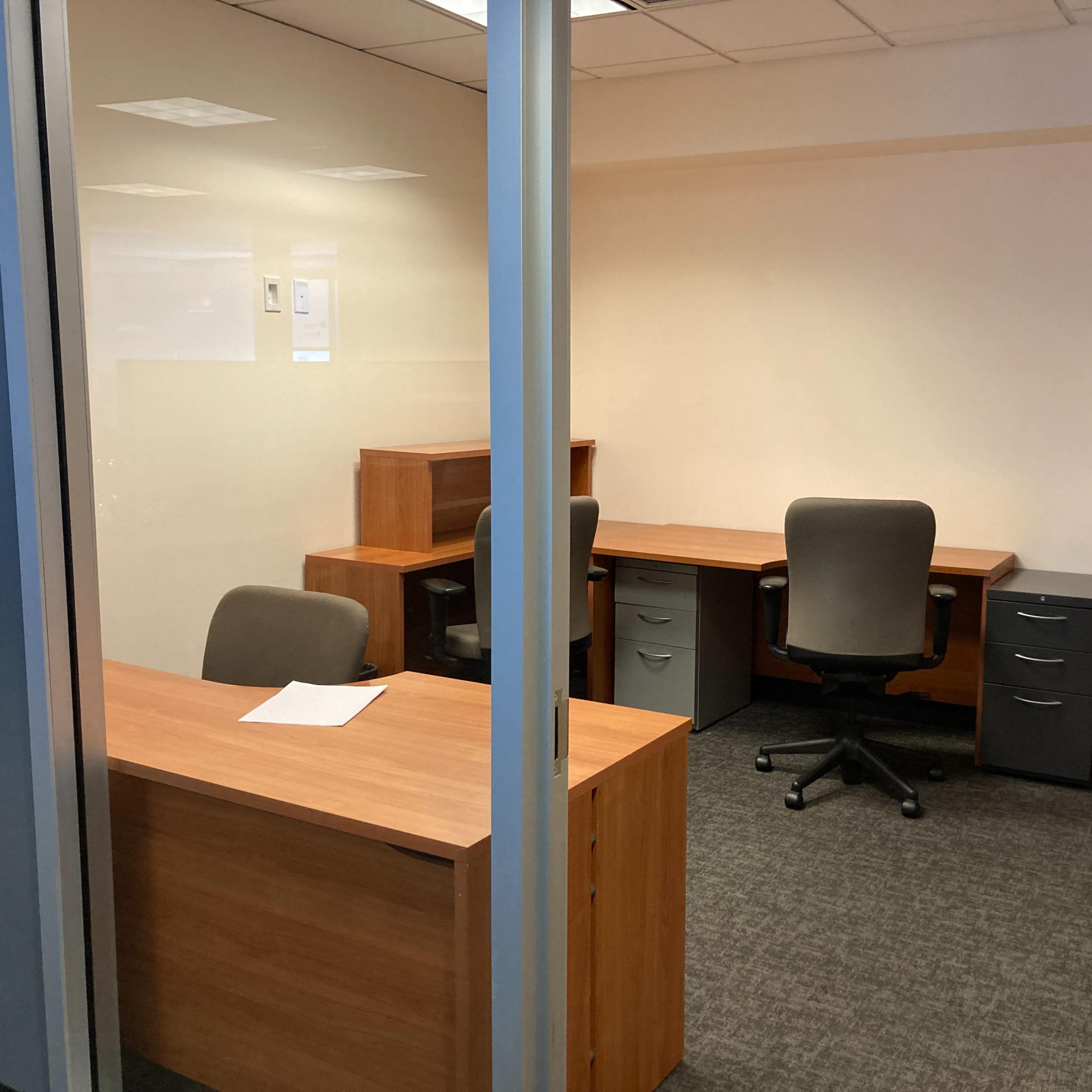A simple office space with two wooden desks, grey filing cabinets, and chairs, all situated within a glass-walled room.