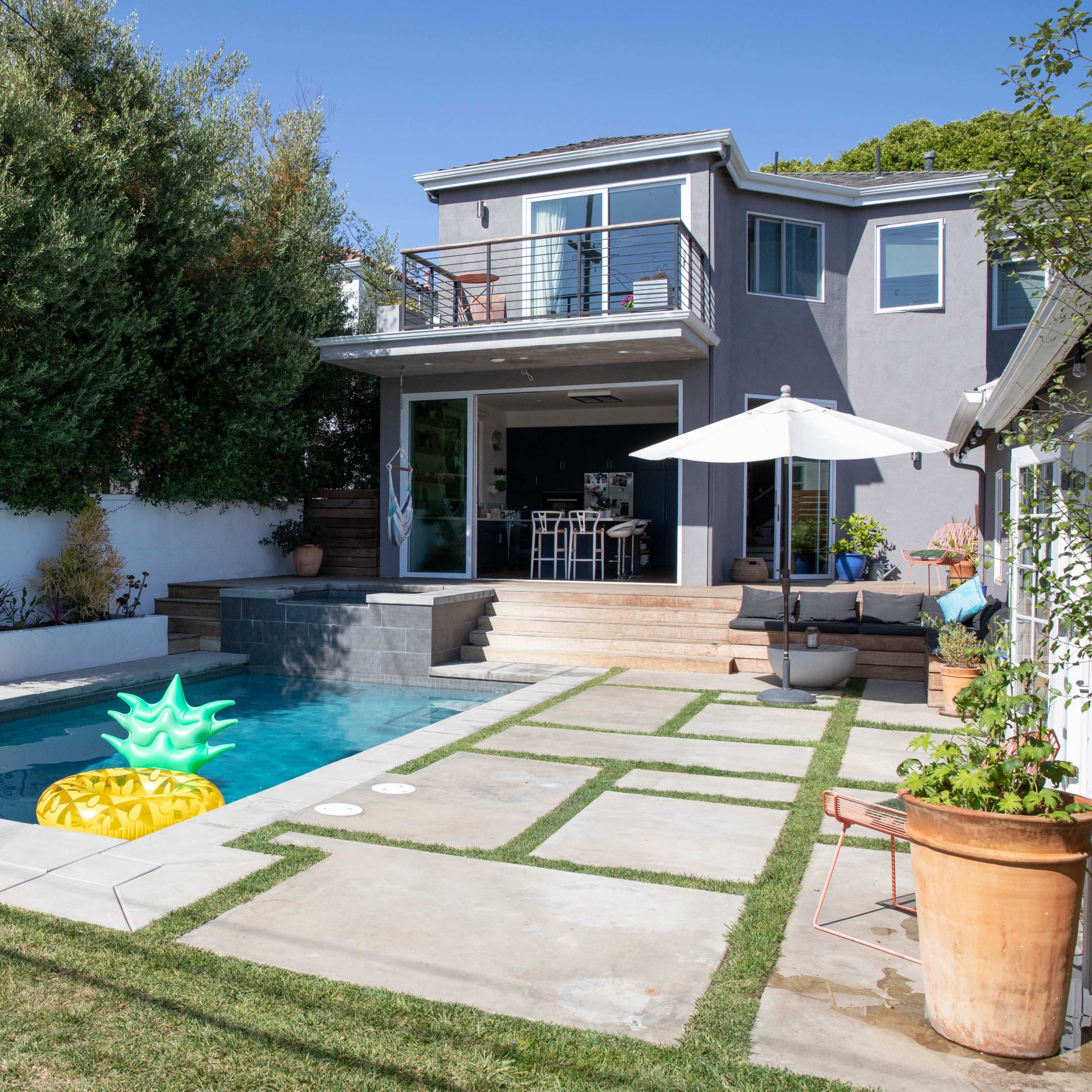 A modern backyard featuring a pool with a pineapple float, surrounded by concrete walkways, grass, and a patio with outdoor furniture.
