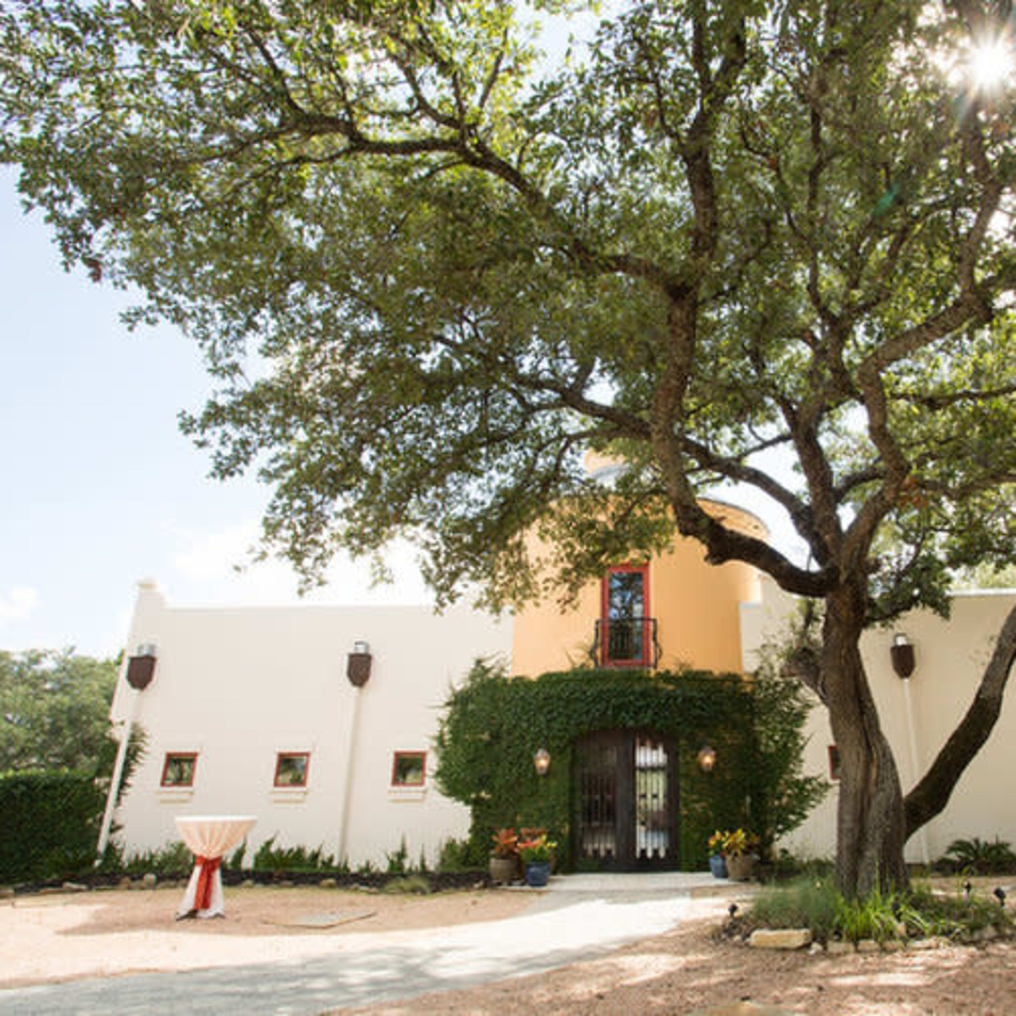 A white building with orange accents is surrounded by large trees and has outdoor tables set up nearby.
