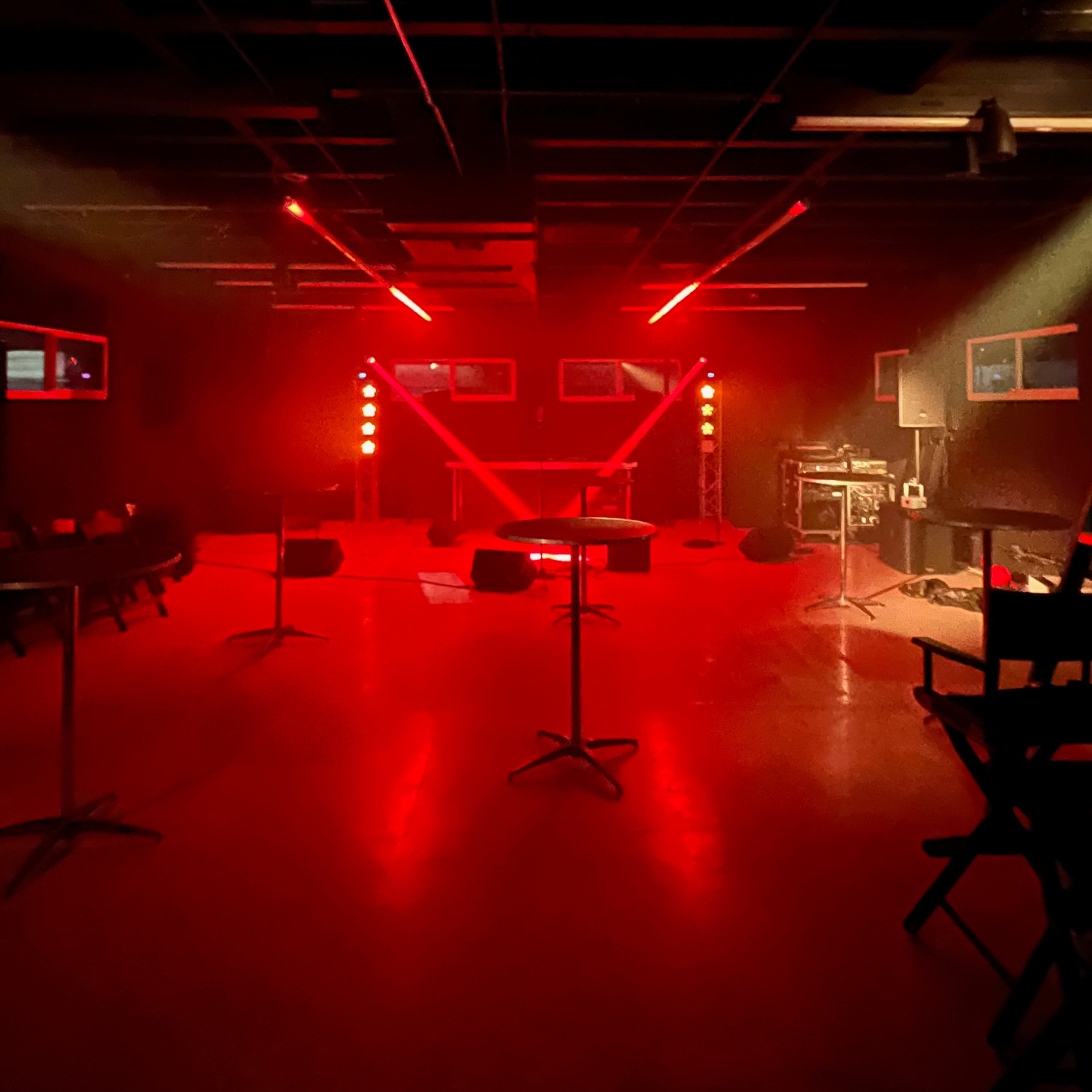 A dimly lit performance space featuring red stage lights, empty seating, and equipment positioned for a live event.