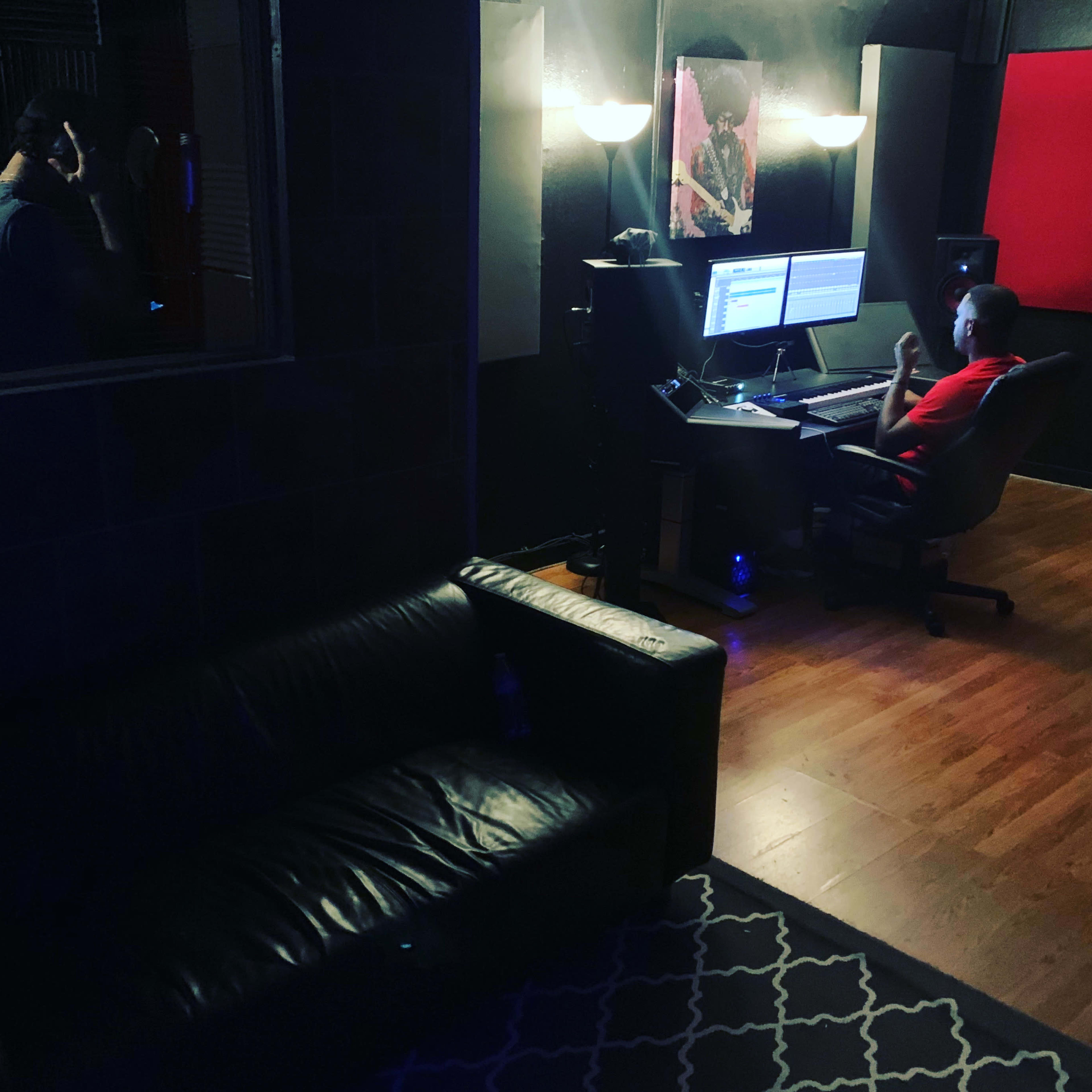 A person sits at a computer desk in a recording studio, while another stands in a separate area, both surrounded by soundproofing panels.