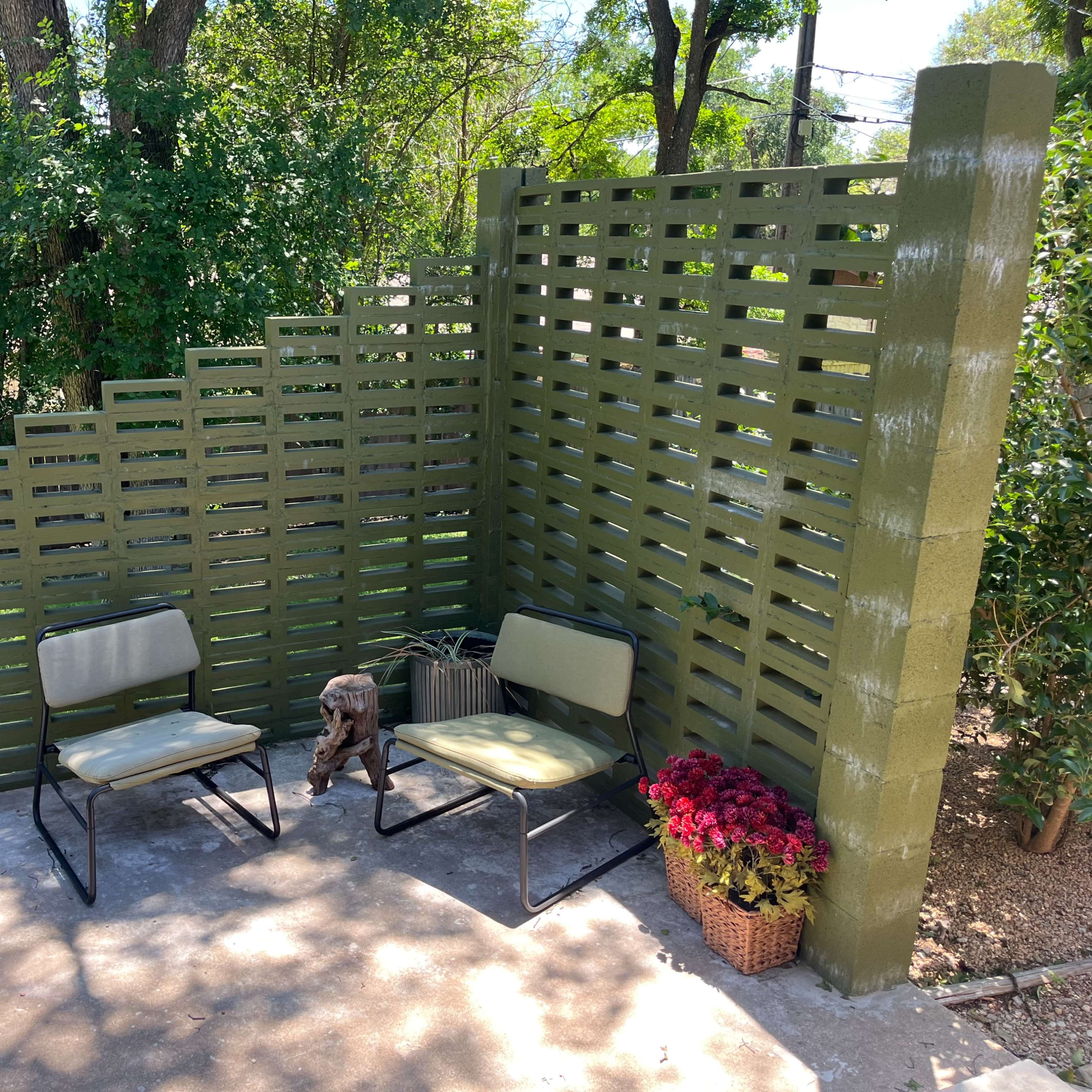 A small outdoor seating area features two metal chairs, a rustic side table, and potted flowers against a vertically slatted green wall.