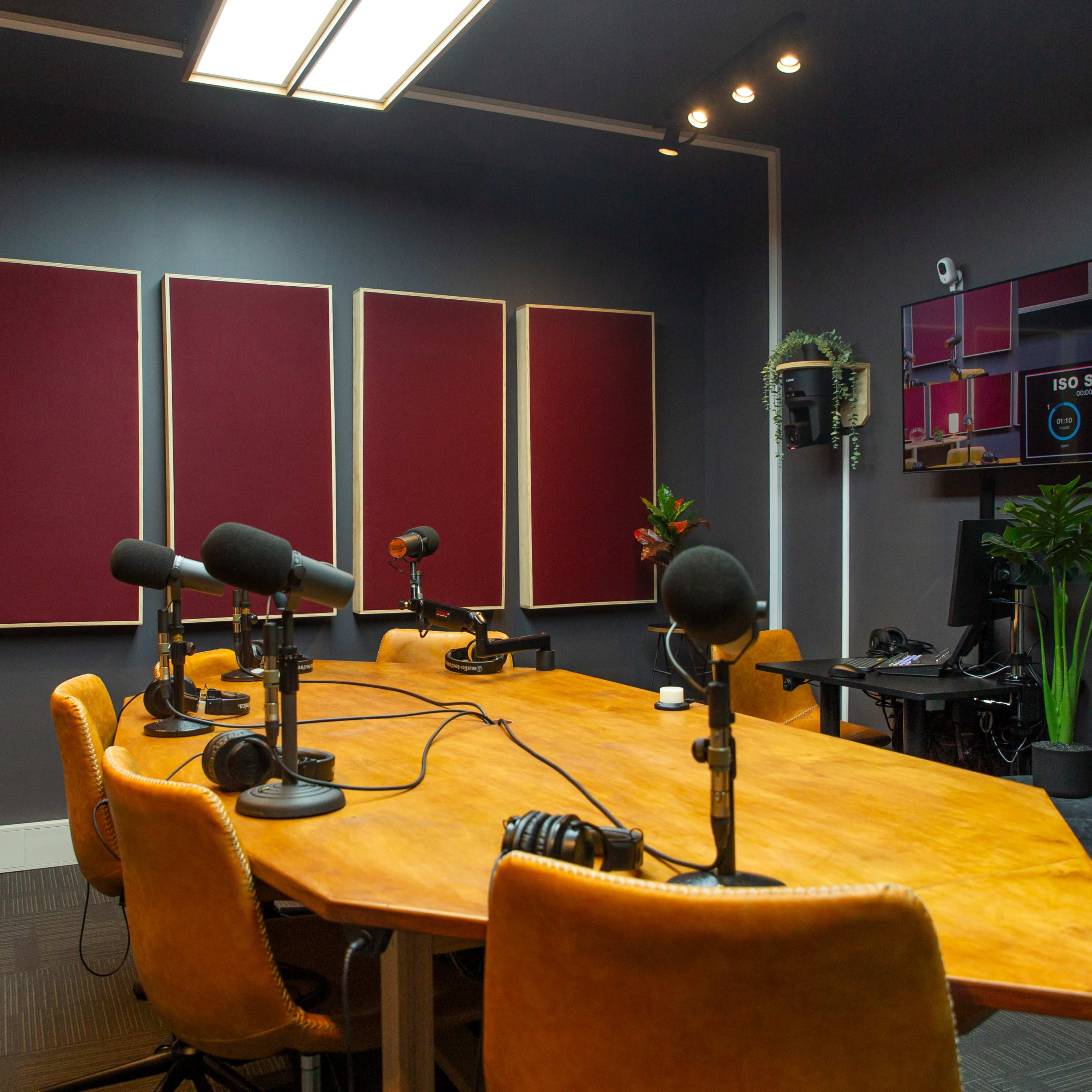 A modern podcast studio features a large wooden table surrounded by several microphones and chairs, with soundproofing panels on the walls and a monitor displaying control options.