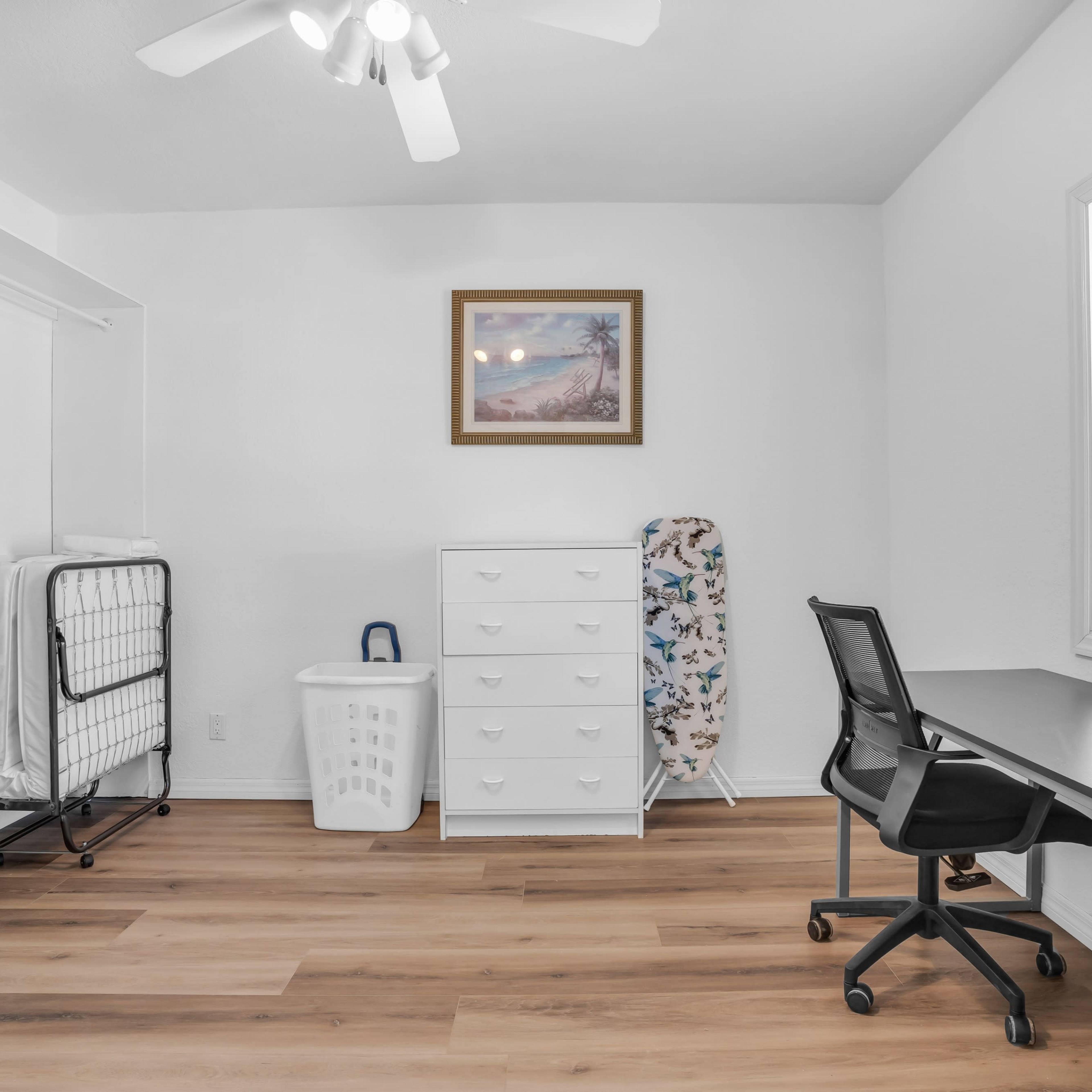 A sparsely furnished room with a white dresser, a folding laundry rack, an ironing board, a laundry basket, and a desk with a chair under a ceiling fan.