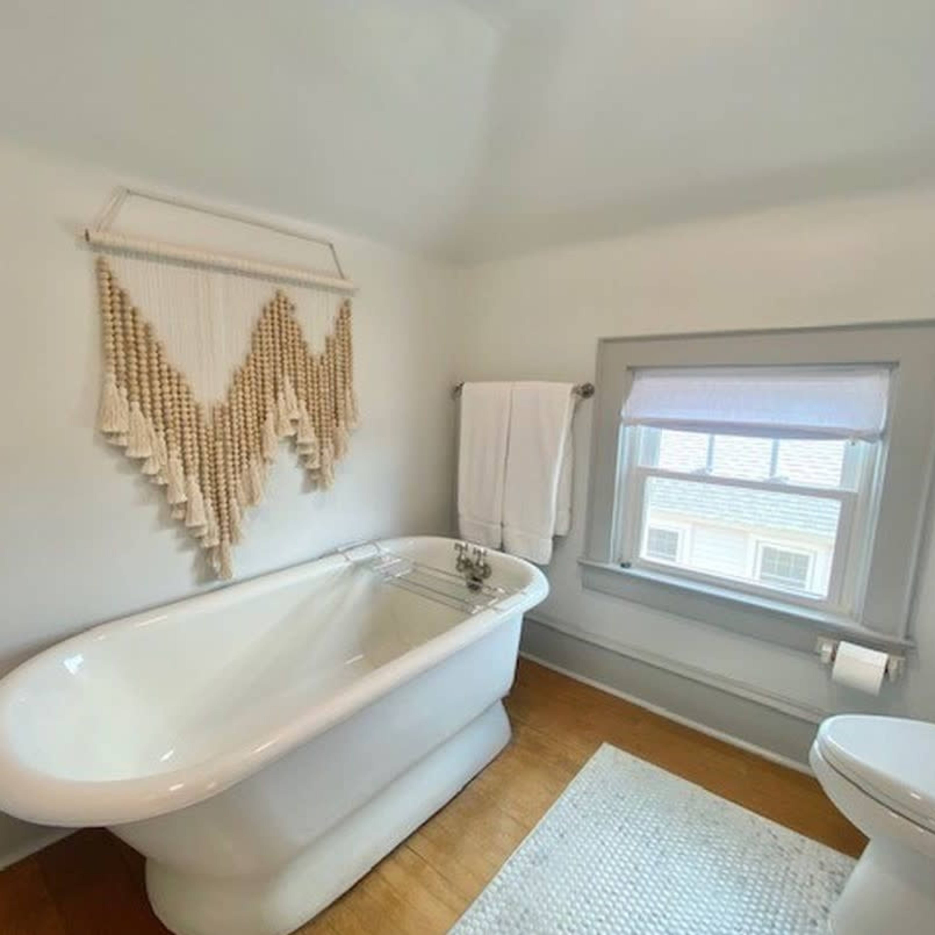 A bathroom featuring a white freestanding bathtub, a white towel hanging on a rack, a window with a light-colored shade, and a patterned rug on the wooden floor.
