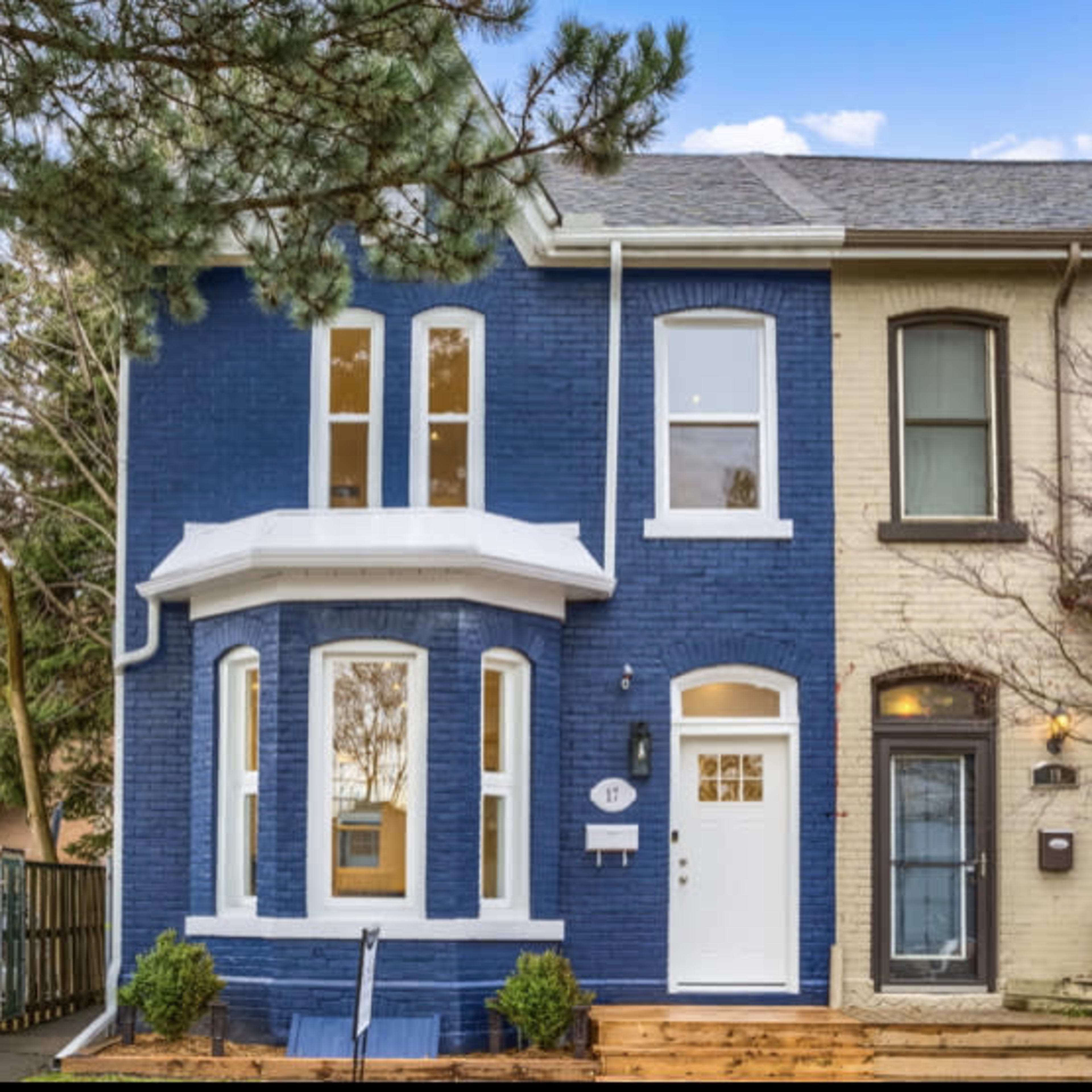 A two-story brick townhouse with a blue facade and white trim, situated next to a light-colored neighboring building and featuring a wooden front porch.