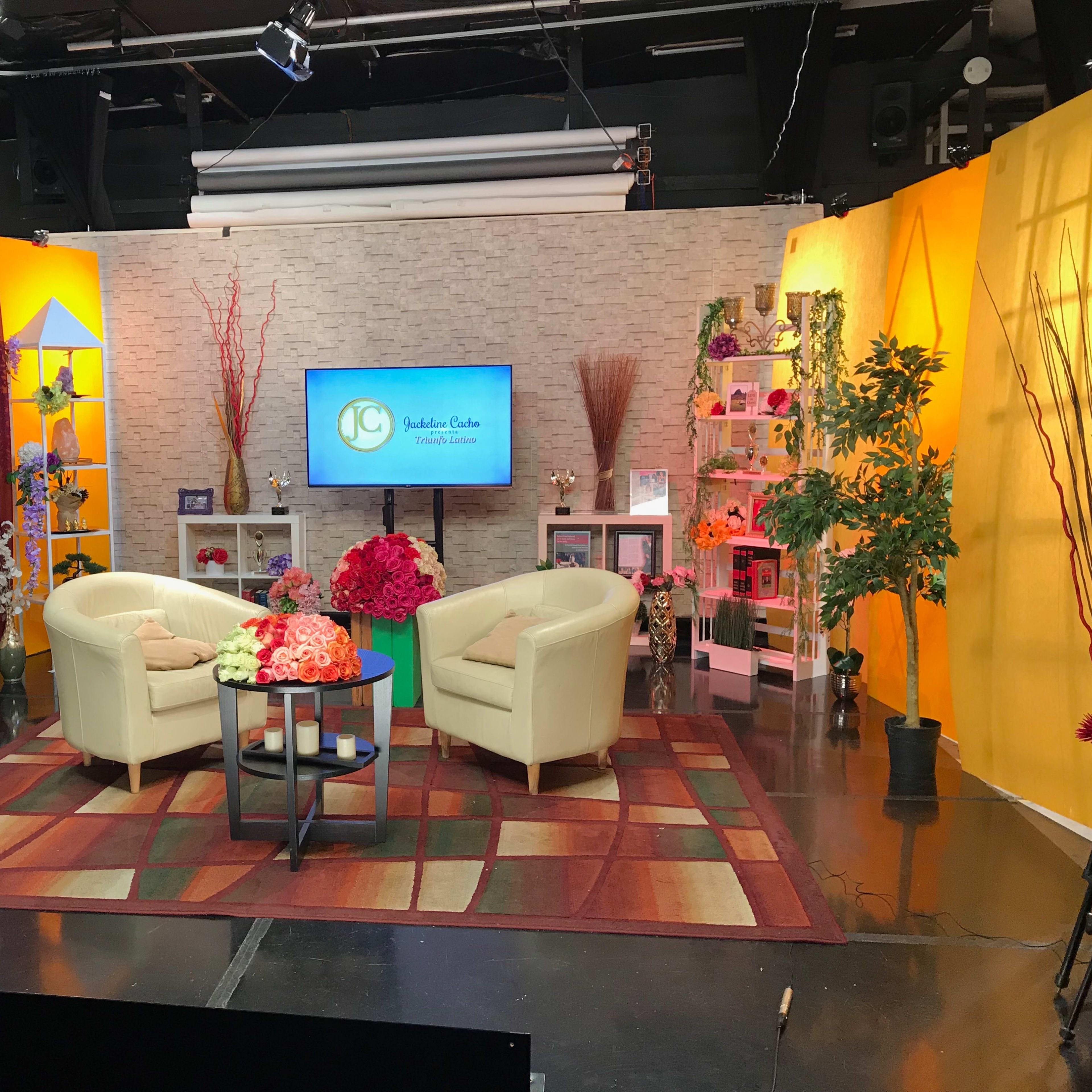 A television studio set featuring two cream-colored chairs and a round table, surrounded by colorful decorations and potted plants.