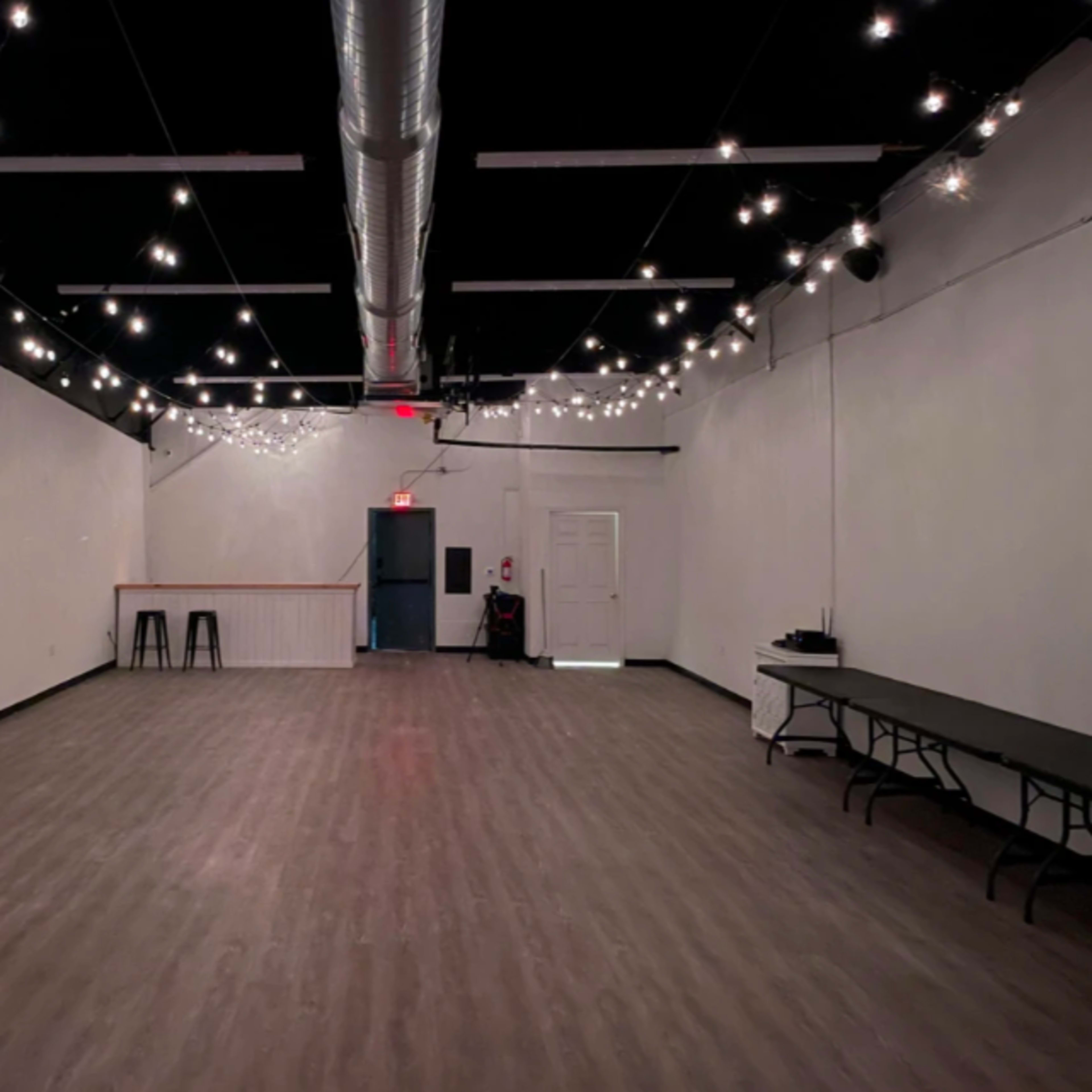 An empty indoor space with wooden flooring, white walls, and string lights overhead.