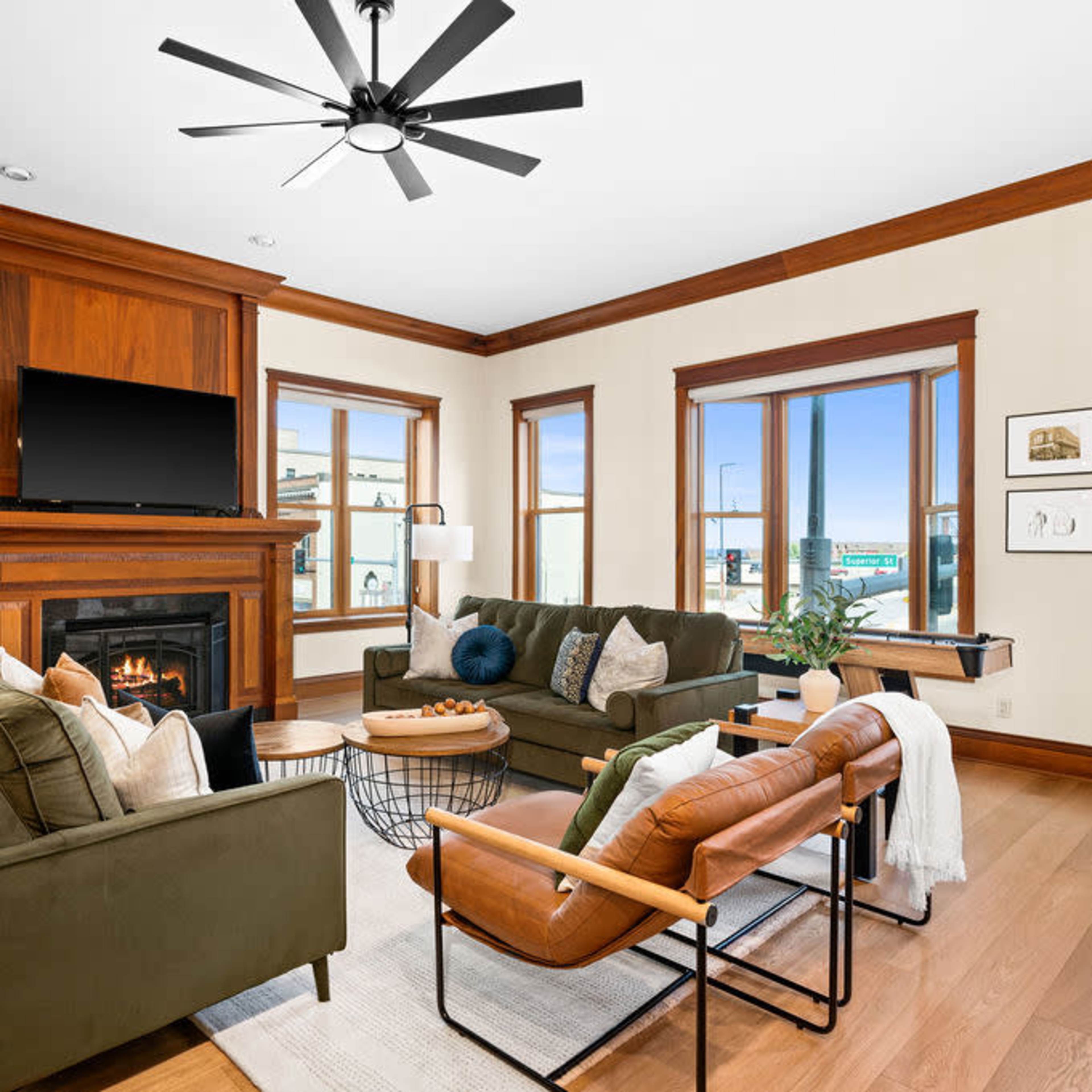 A living room with a green sofa, an armchair, a fireplace with a TV above it, and large windows providing natural light.
