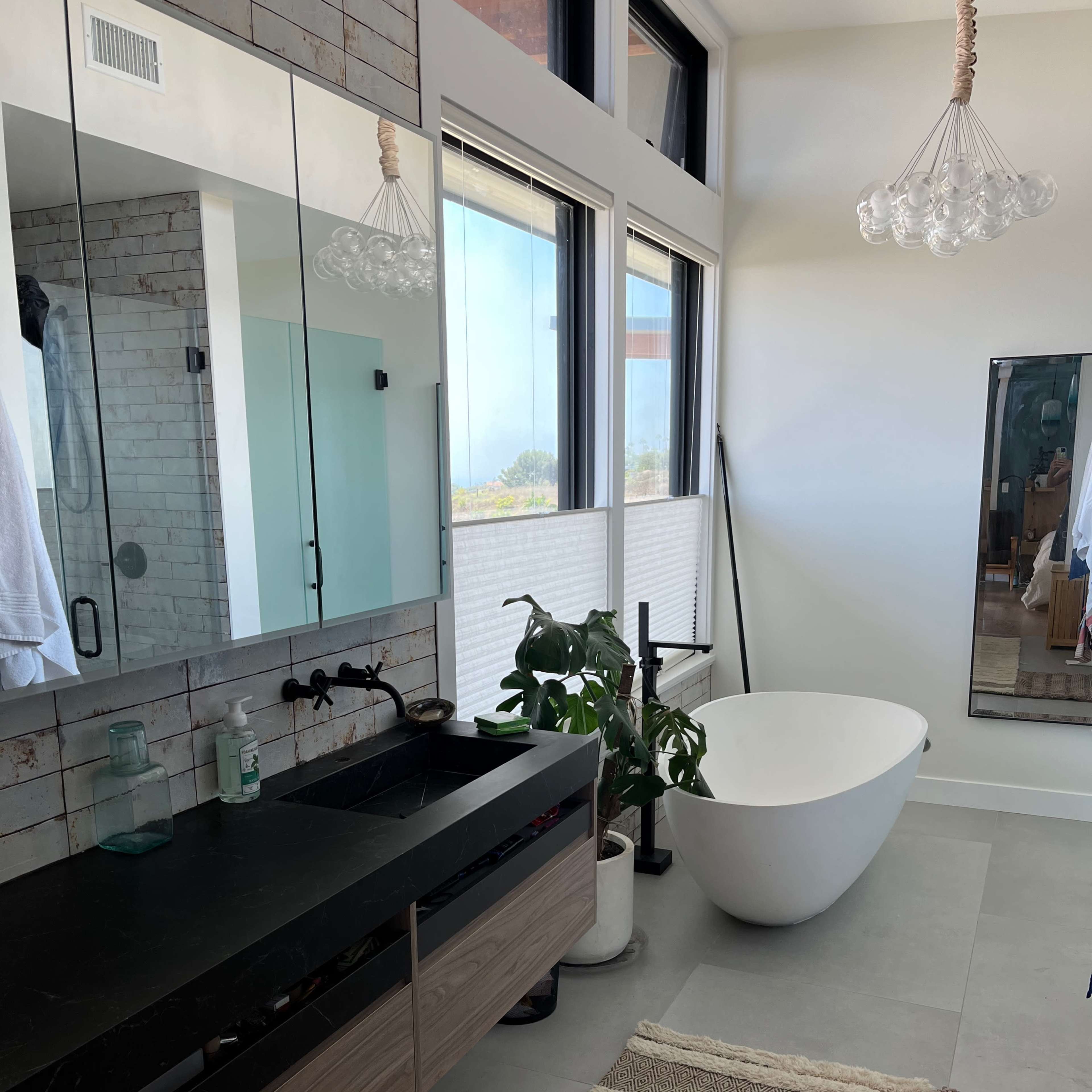 A modern bathroom featuring a freestanding bathtub, a large mirror above a dark sink vanity, and large windows allowing natural light.