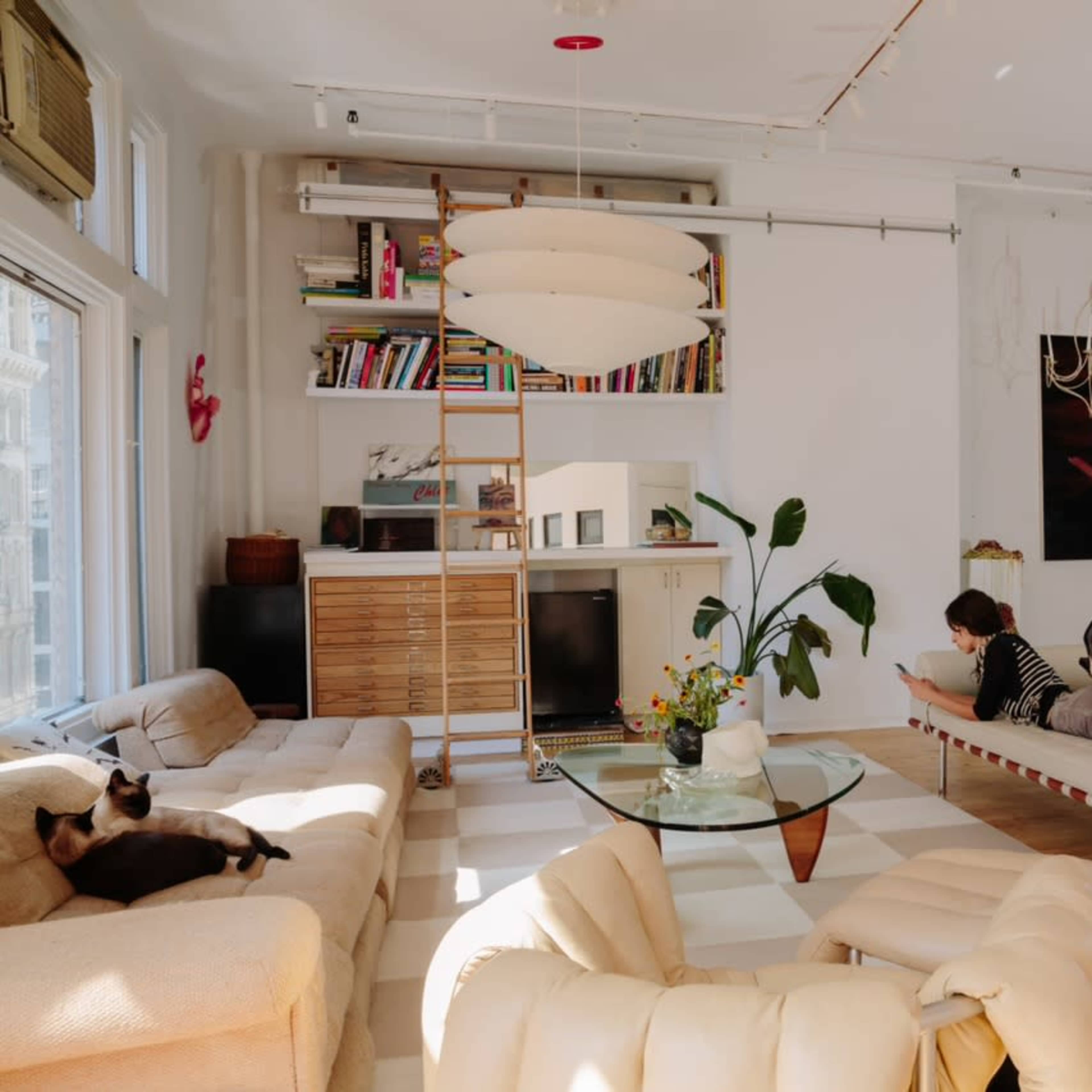 A spacious living room features a light-colored couch, a minimalist seating area, and two large artworks on the wall, while a person relaxes on a daybed and a cat rests nearby.