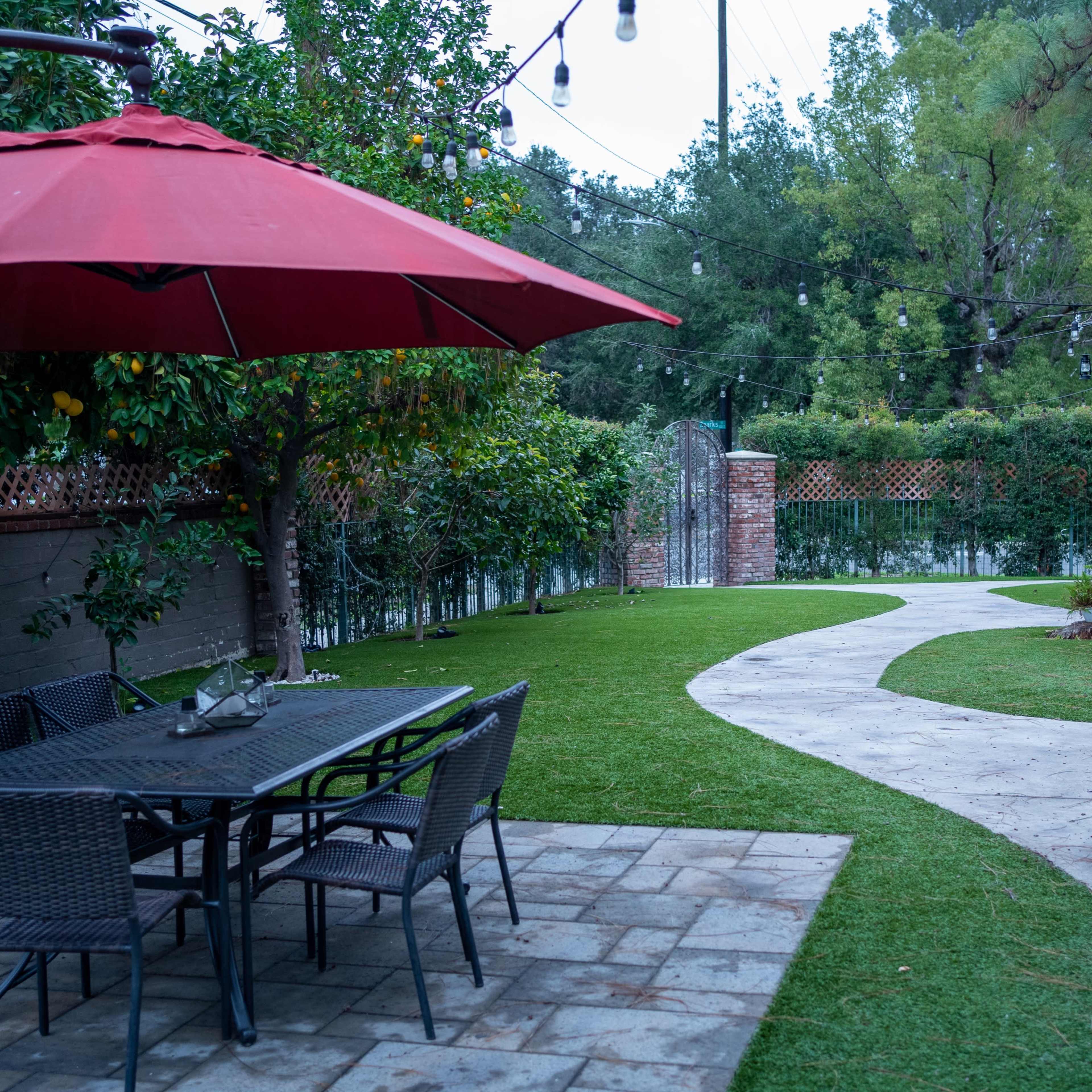 A landscaped garden with a curved pathway, a dining set under a red umbrella, and greenery along the fence.