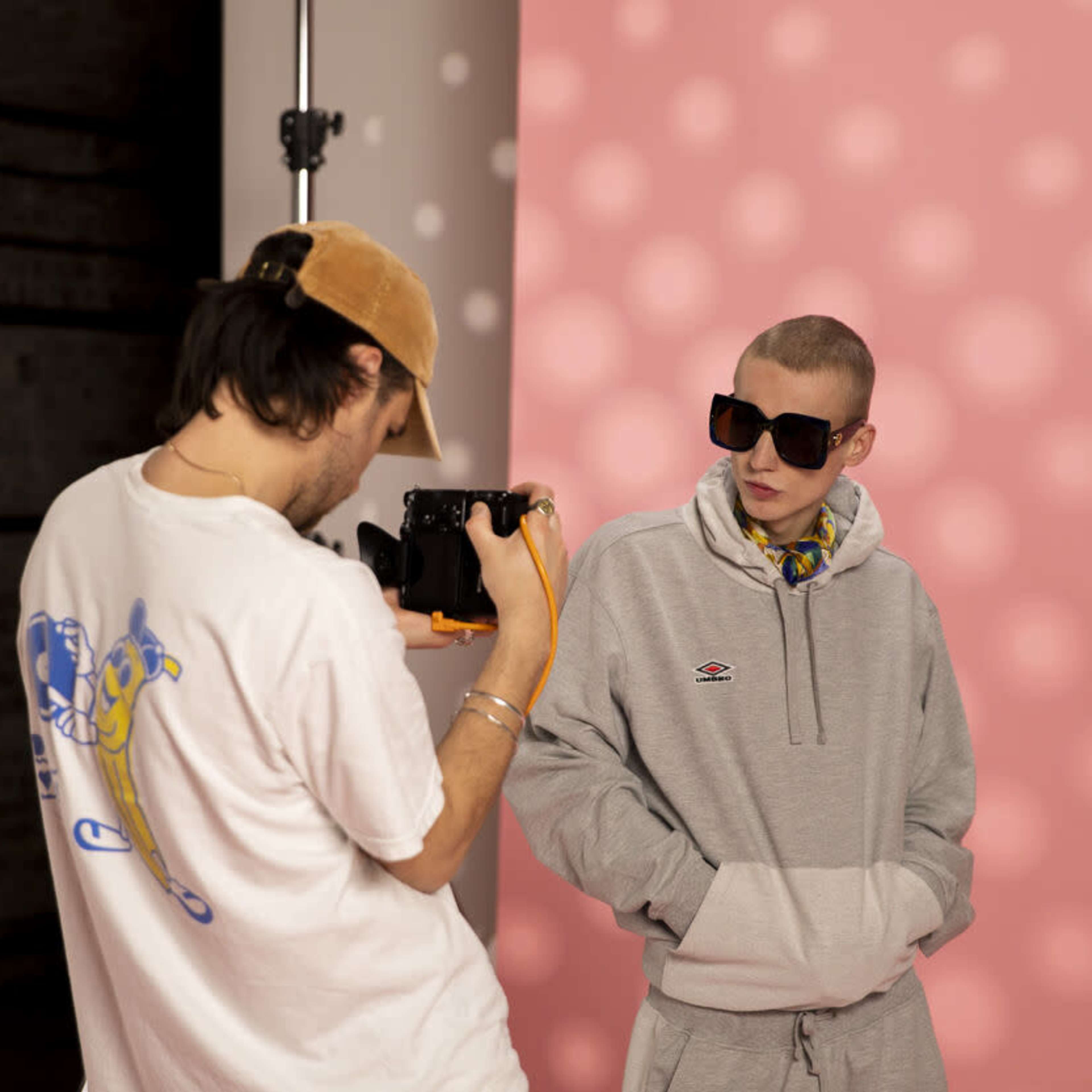 A photographer takes a picture of a model wearing a gray hoodie and sunglasses against a pink backdrop.