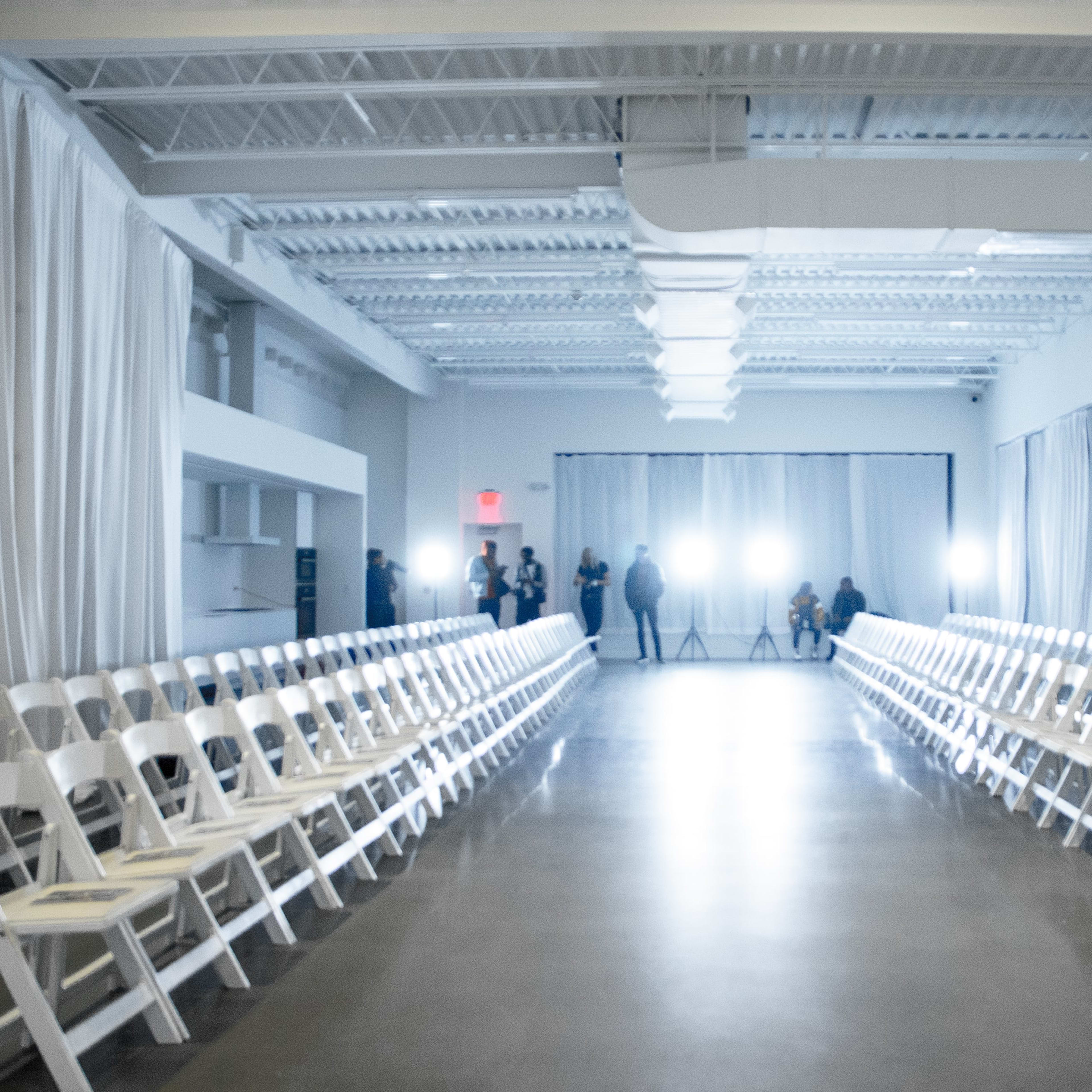 A spacious, well-lit room features rows of white folding chairs facing a central runway with bright lights illuminating the space.