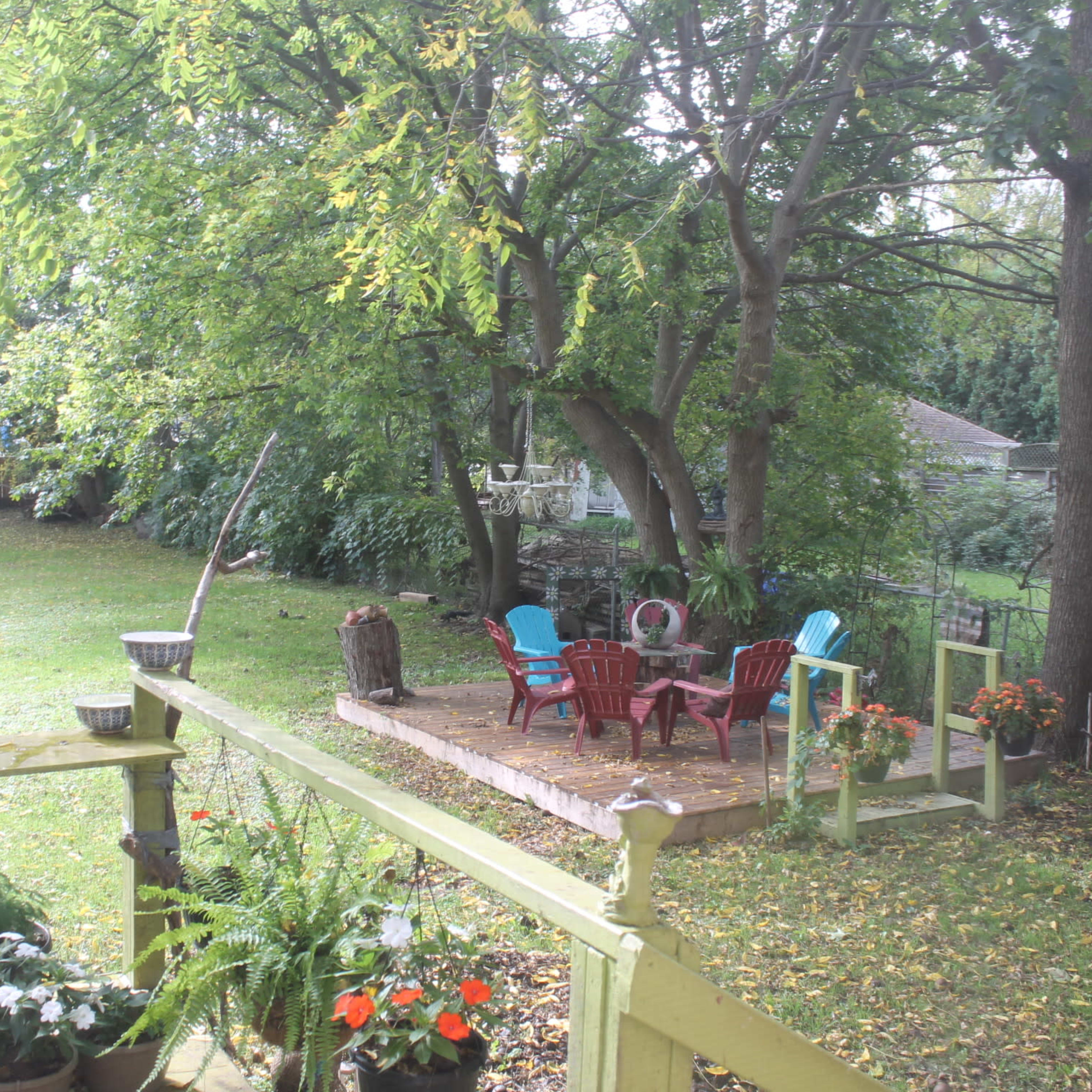 A spacious backyard with a patio area featuring colorful chairs surrounded by trees and garden plants.