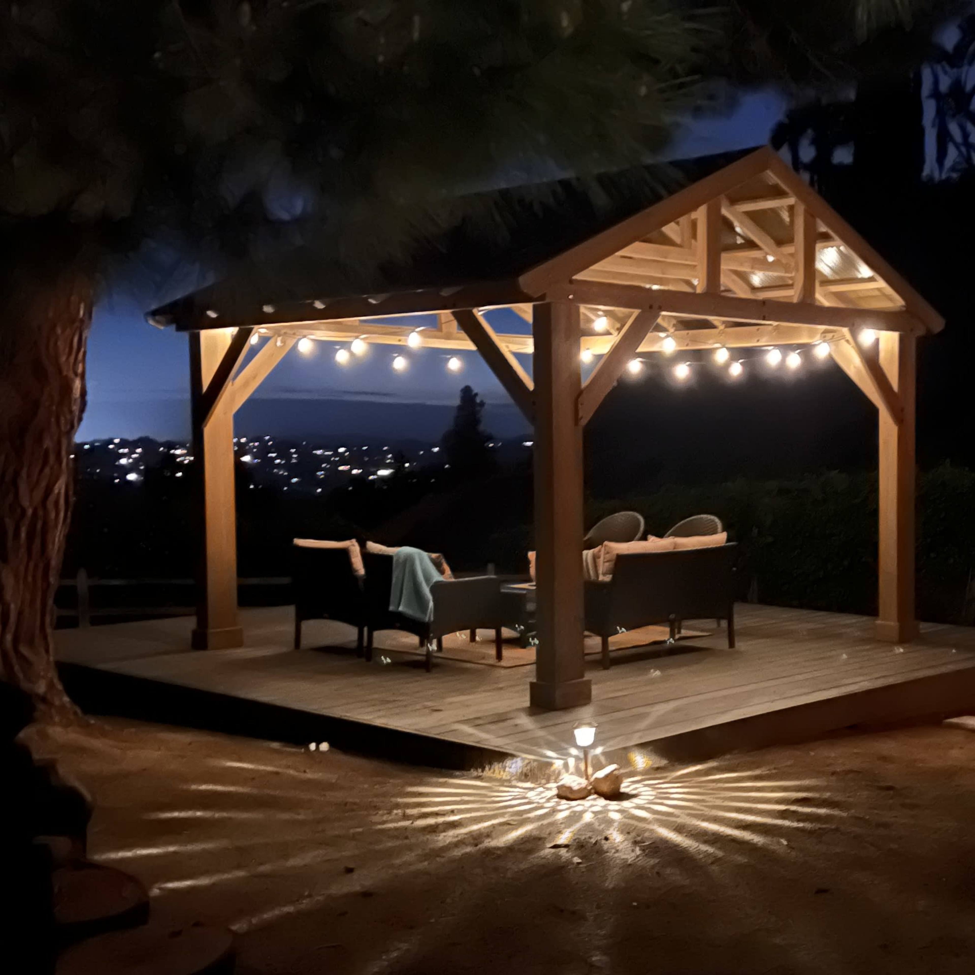 A wooden gazebo with string lights stands on a deck, surrounded by a softly lit garden at night.