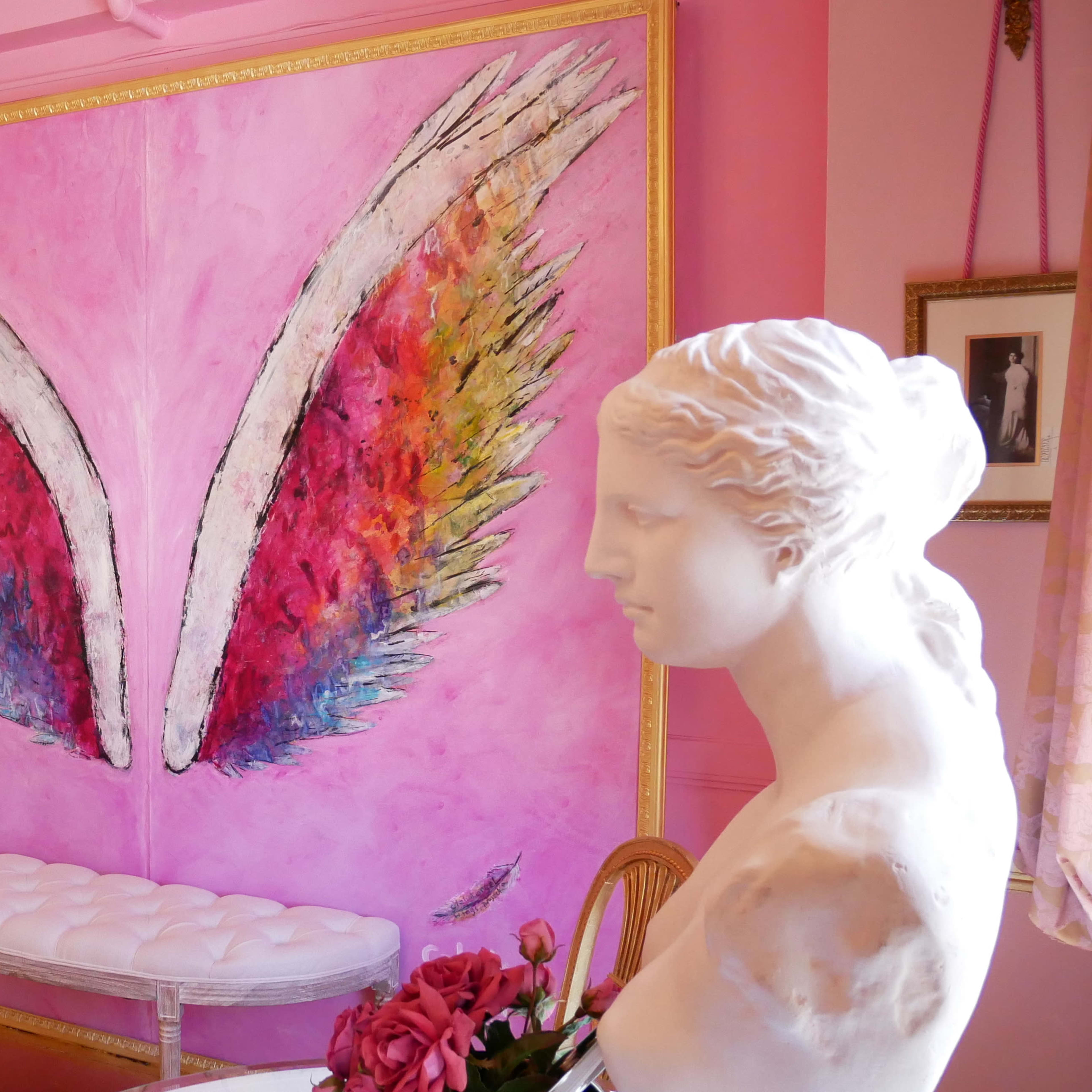 A white statue of a woman is positioned beside a large pink wall mural featuring colorful wings.