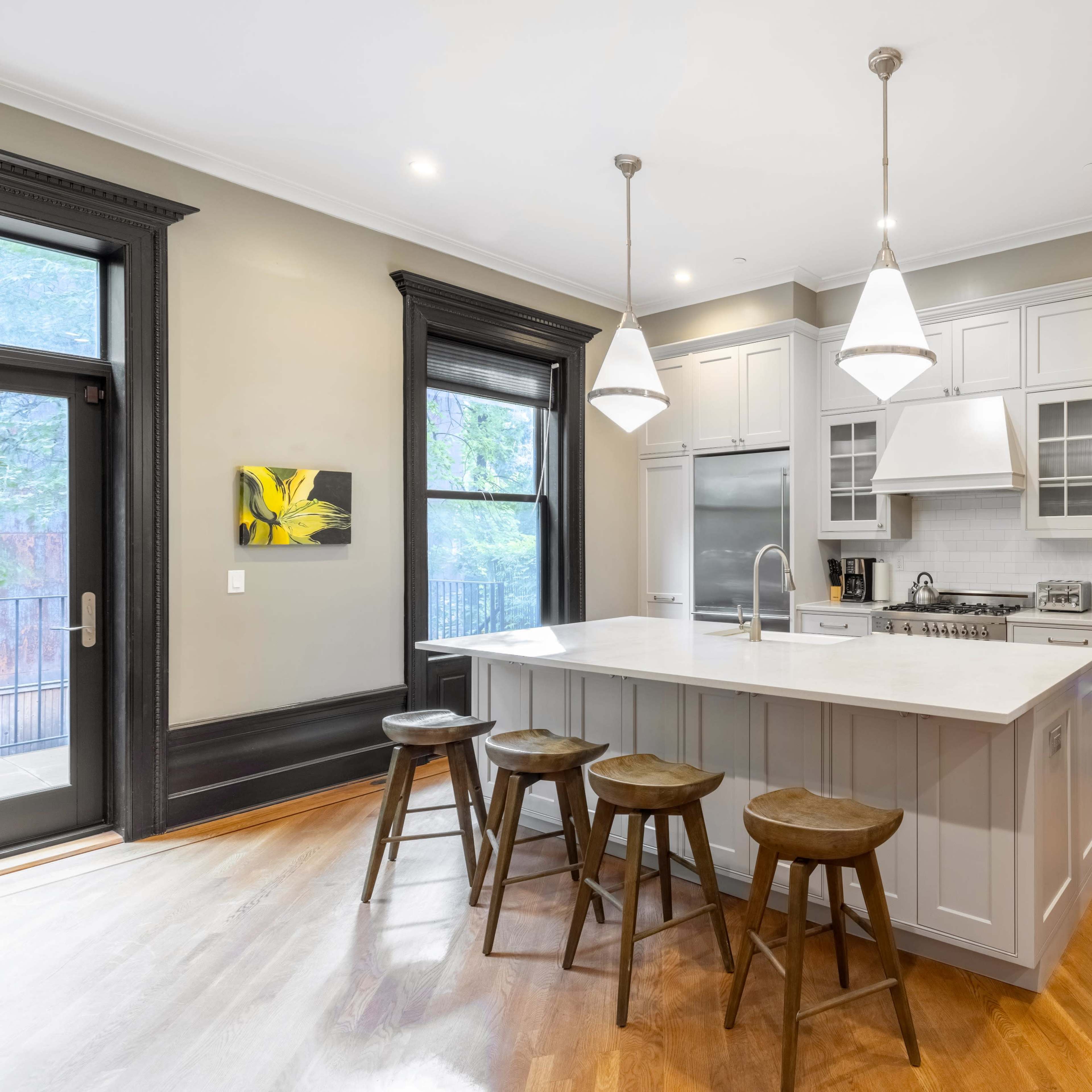 A modern kitchen features a large island with four wooden stools, gray cabinetry, and stainless steel appliances.