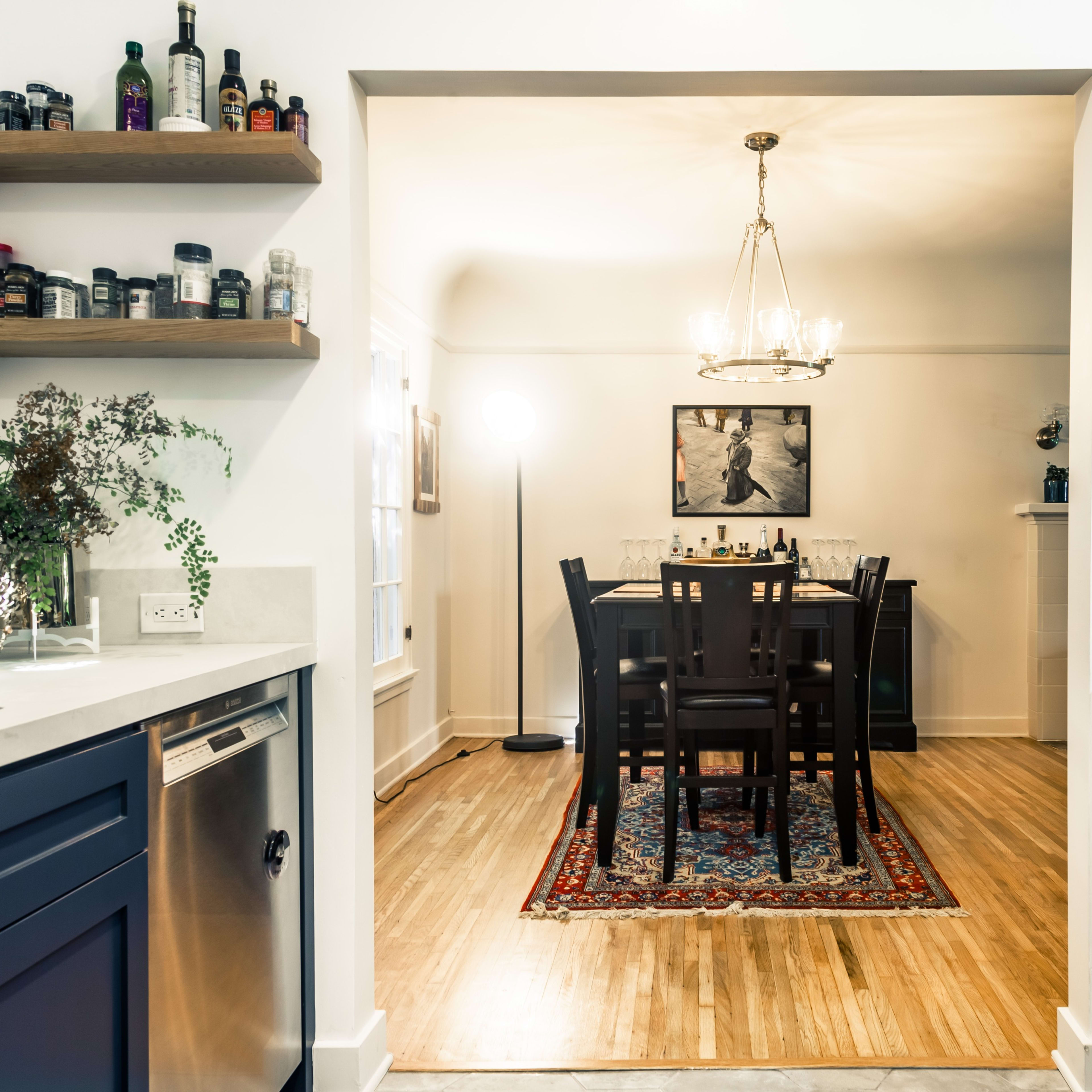 A dining area featuring a table set for four, illuminated by a chandelier, is seen through an open doorway from a kitchen with dark cabinetry and decorative shelves.