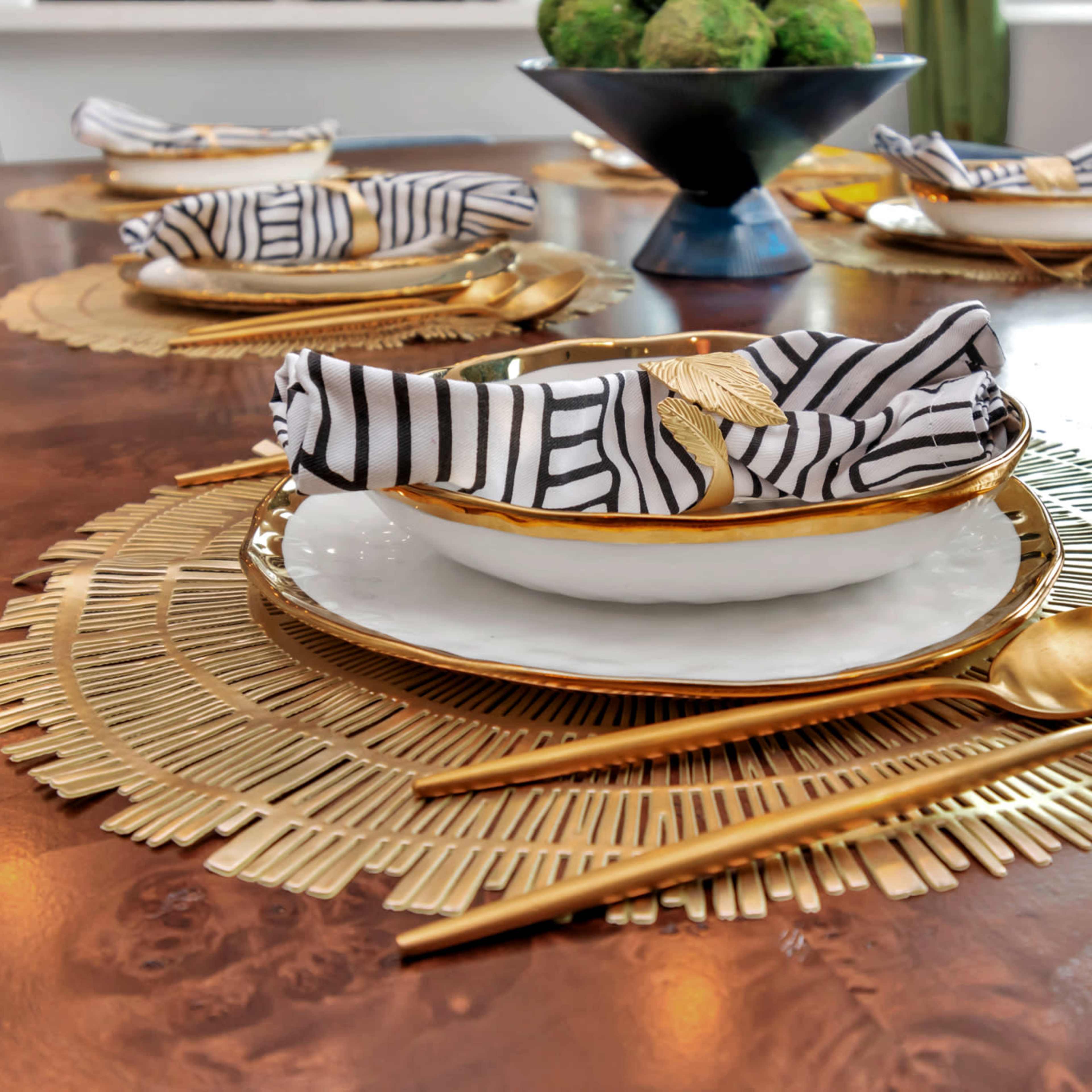 A dining table is set with gold-accented plates, striped napkins, and decorative utensils, surrounded by a textured placemat and a centerpiece.