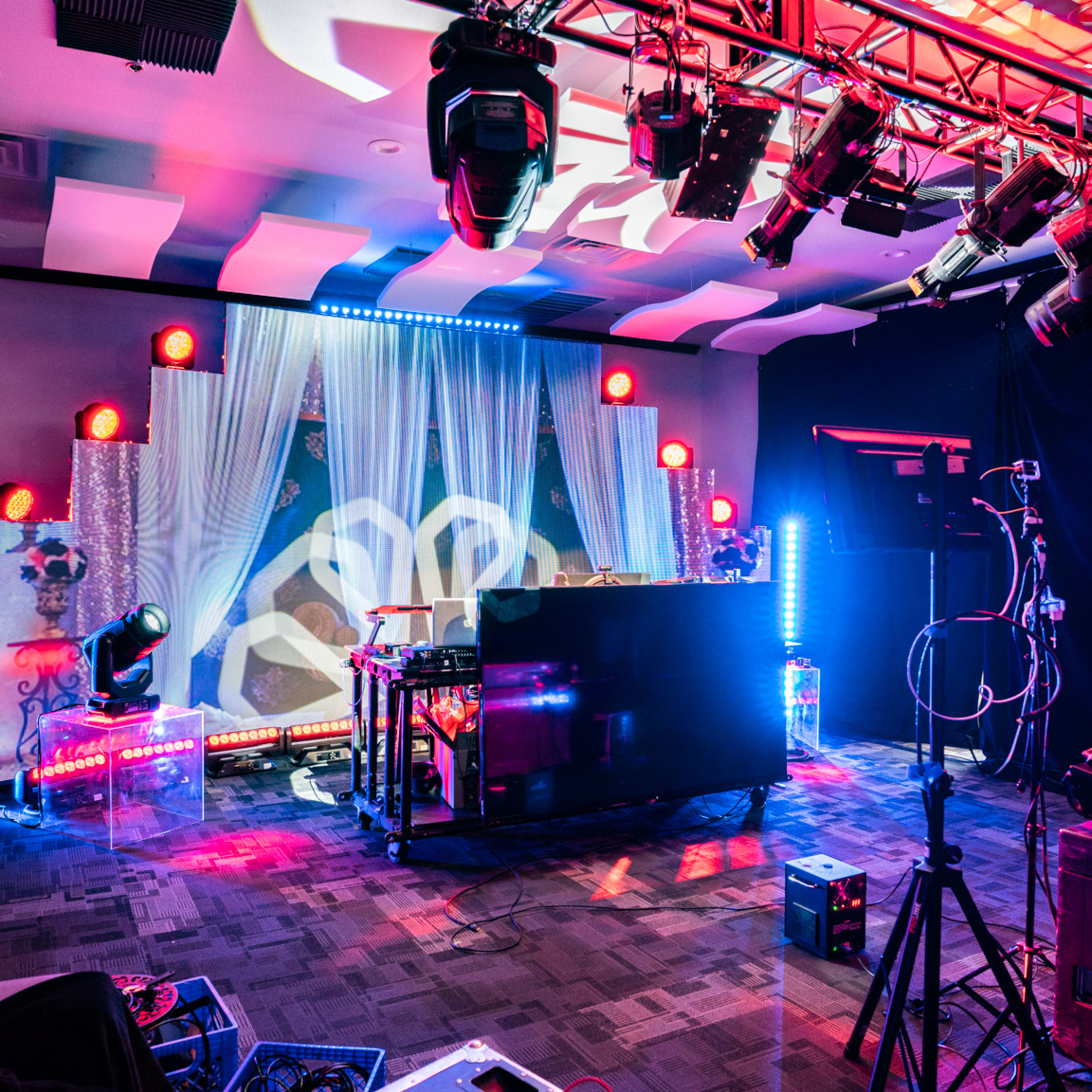 A stage setup featuring a DJ booth with colorful lighting, surrounded by sound equipment and a backdrop of draped fabric.
