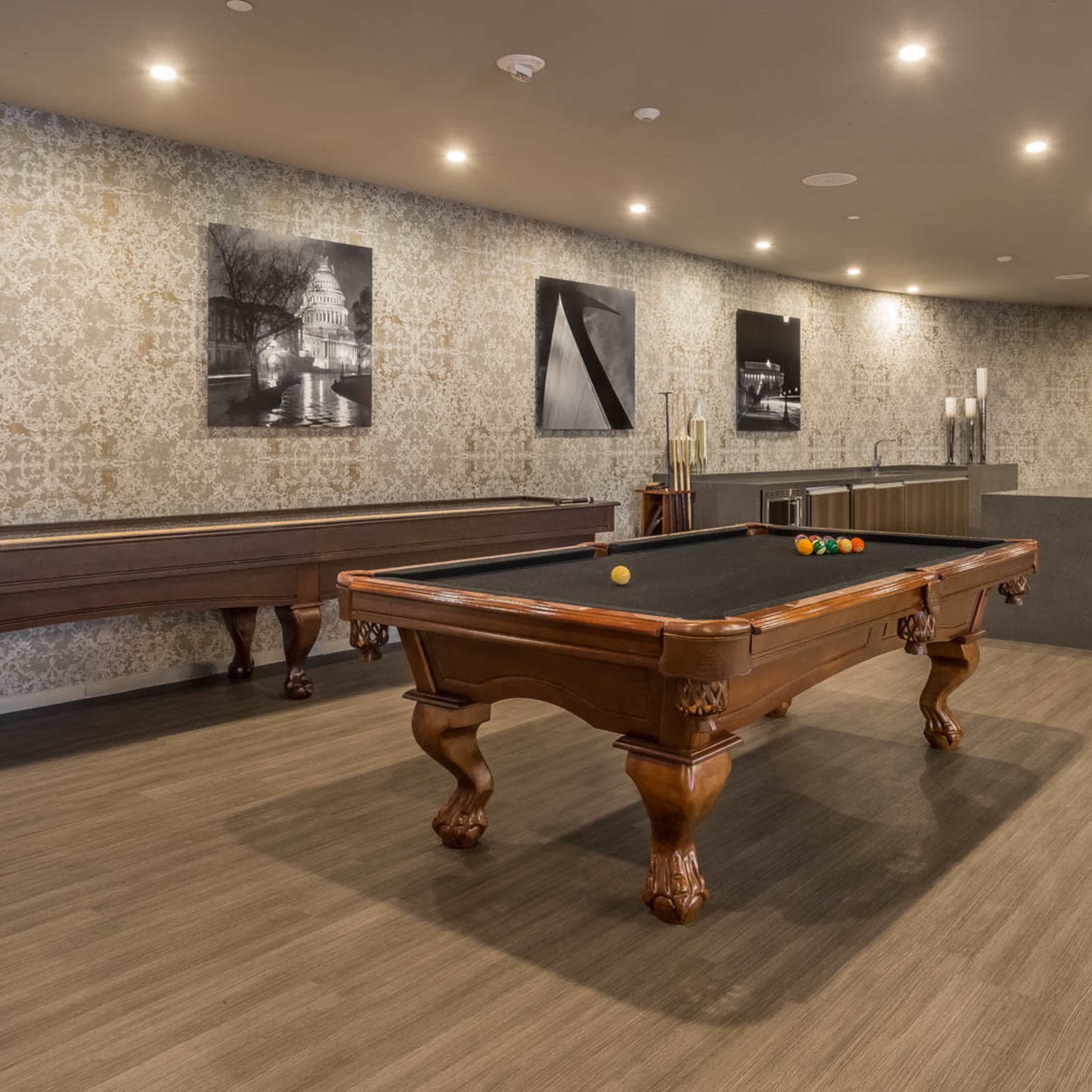 A game room featuring a billiard table, a wooden foosball table, and a bar area with blue stools against a patterned wall.