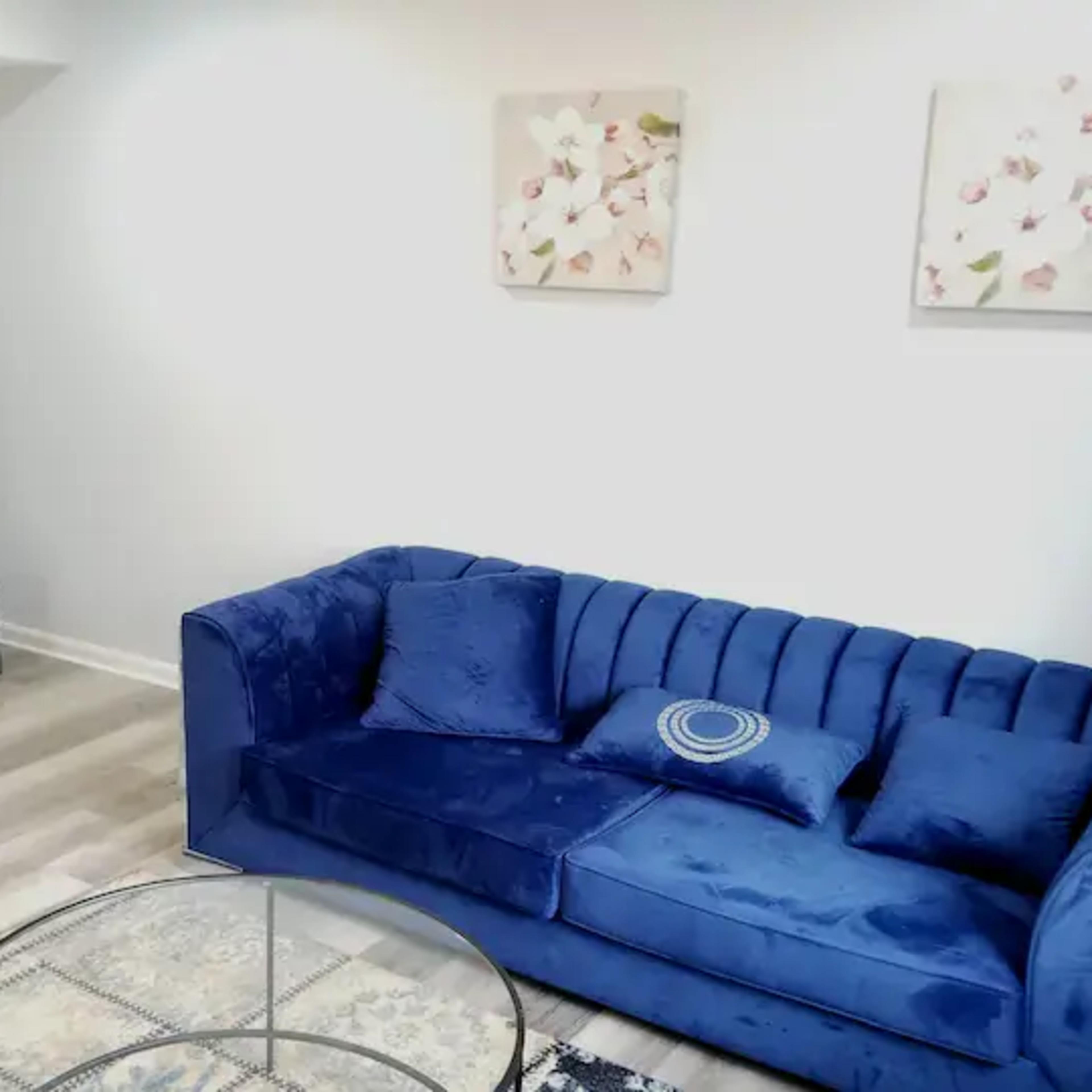 A living room with a blue velvet sofa, a glass coffee table, and a large television on a black stand against a white wall.