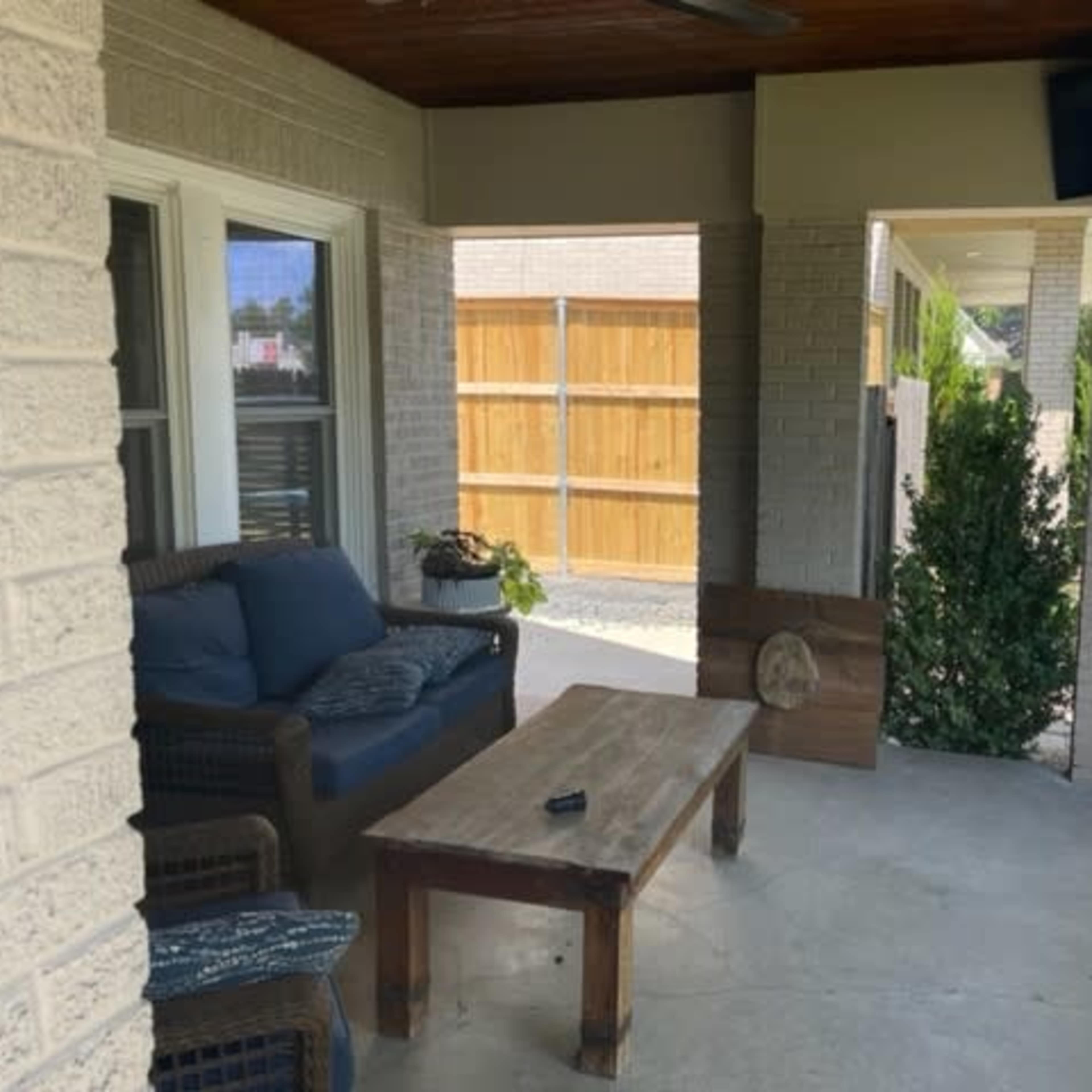A covered porch with a wicker seating area, a wooden coffee table, and a view of a fenced yard.